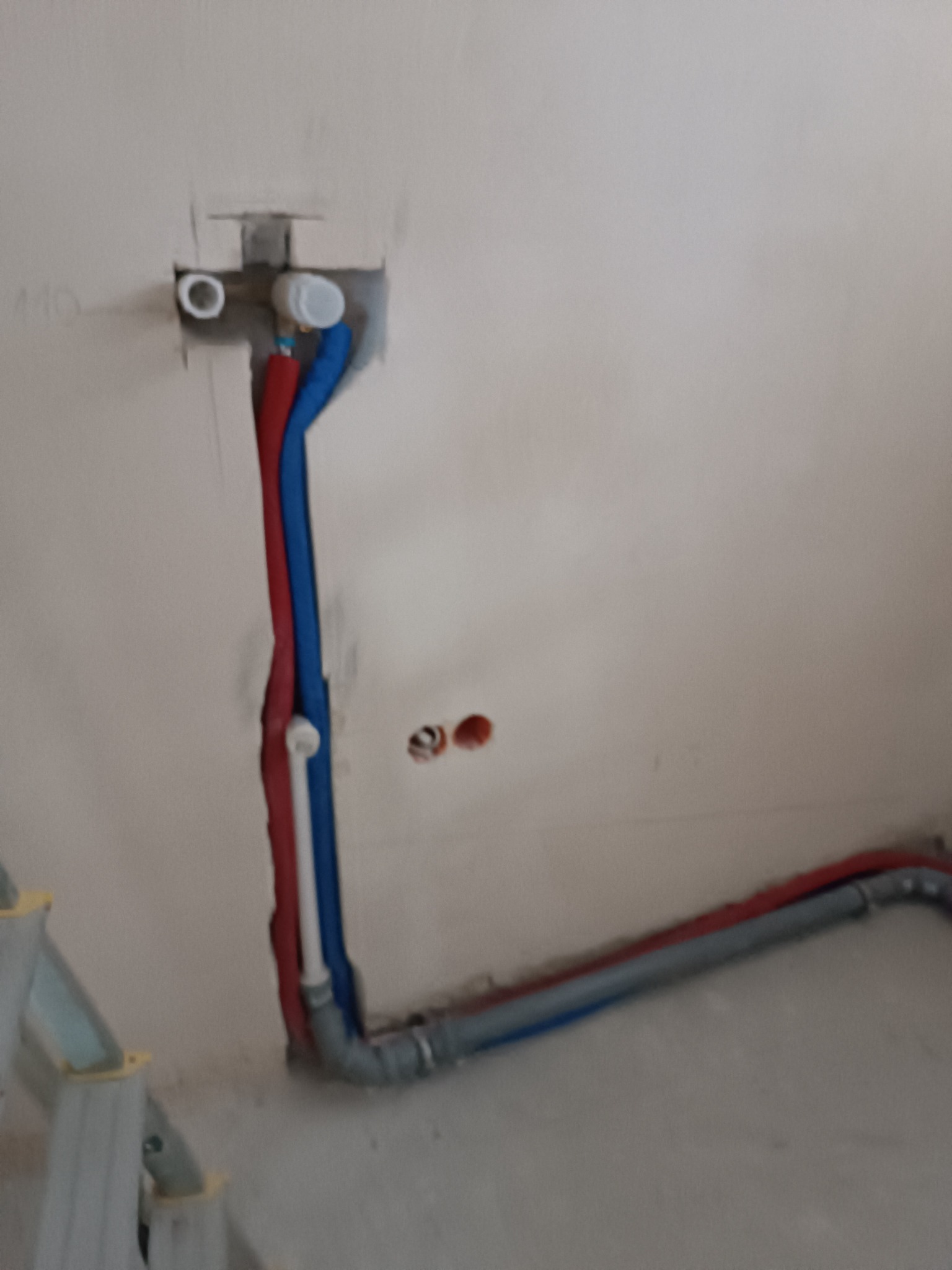 Nowe, częściowo ukryte w ścianie, rury hydrauliczne (czerwone, niebieskie i szare) w trakcie montażu w łazience. Widoczny fragment drabiny.