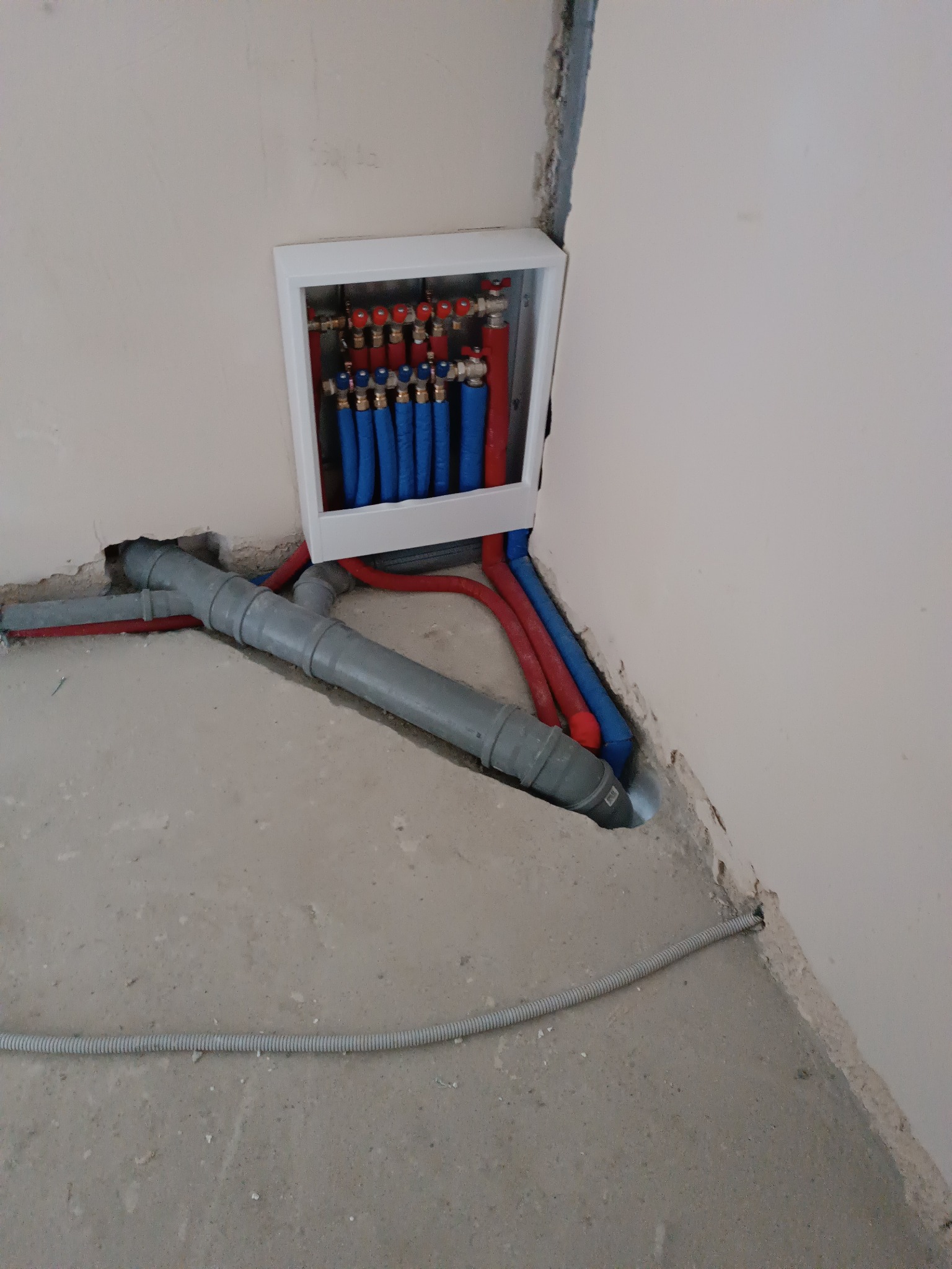 Rozdzielacz ogrzewania podłogowego z niebieskimi i czerwonymi rurami w białej obudowie, szare rury kanalizacyjne na surowej podłodze. Instalacja w trakcie realizacji.