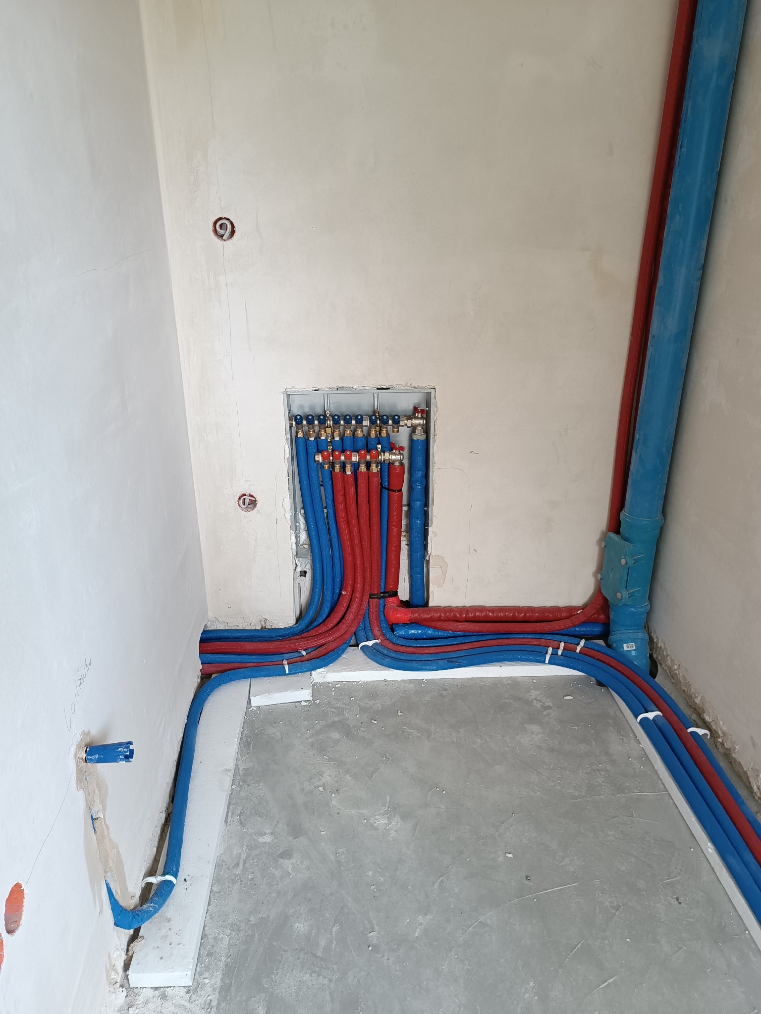 Instalacja rur w kolorze niebieskim i czerwonym, zamocowanych do ściany i podłogi za pomocą uchwytów. Widoczne przyłącza i otwory w ścianie.