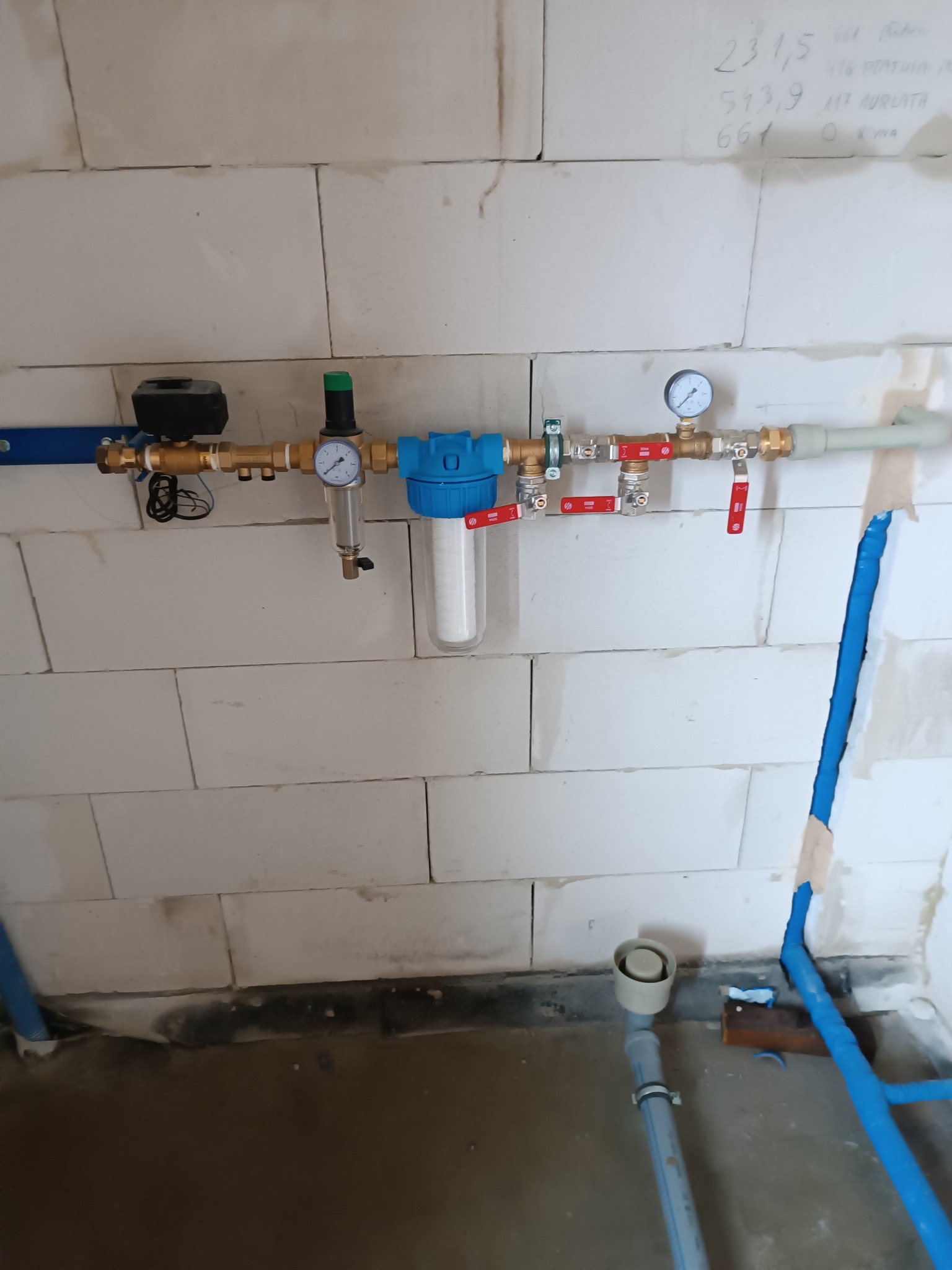 Szczegółowe ujęcie instalacji hydraulicznej z filtrem wody, manometrami i zaworami, zamontowanej na ścianie z białych bloczków betonowych. Widoczne rury i elementy złączne.