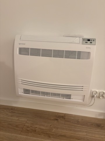 Biały klimakonwektor przypodłogowy w Miedźnej, zamontowany na ścianie. Widoczne kratki wentylacyjne i panel sterowania. Gniazdka elektryczne w pobliżu.