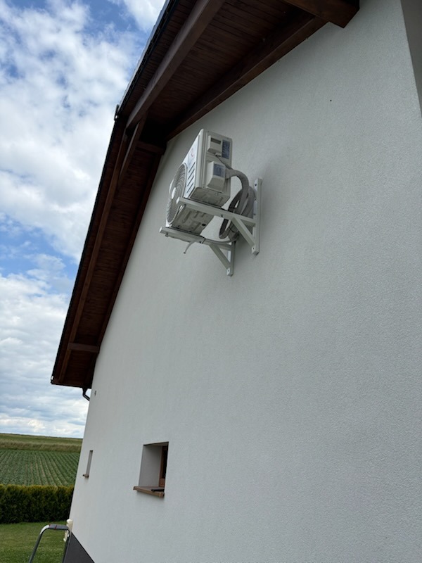 Biała jednostka zewnętrzna klimatyzacji zamontowana na ścianie domu z jasną elewacją. Widoczne elementy montażowe i przewody. W tle zieleń i błękitne niebo.