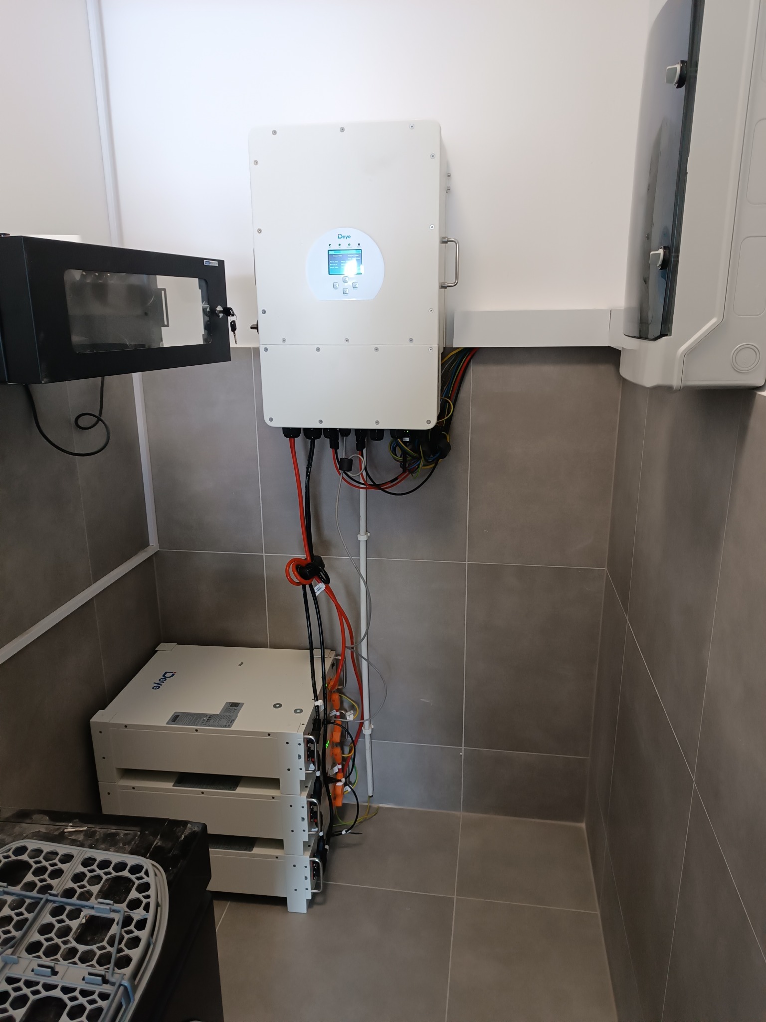 Zamontowany inwerter i baterie fotowoltaiczne Deye na szarej ścianie z płytek. Widoczne przewody i elementy instalacji w pomieszczeniu.