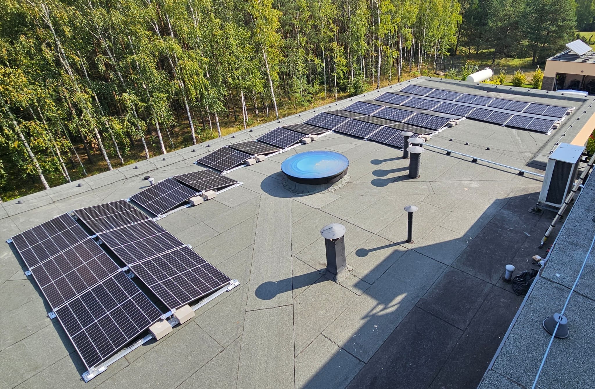 Instalacja paneli fotowoltaicznych na płaskim dachu budynku, widok z góry. Panele ułożone w rzędach, w tle drzewa i fragment budynku. Słoneczny dzień.