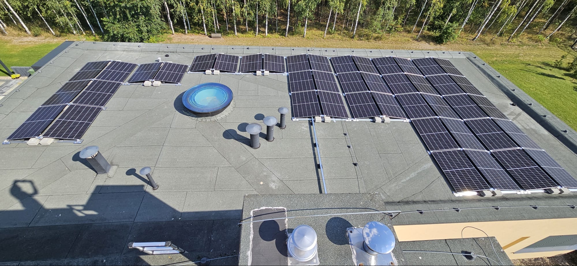 Widok z góry na dach z panelami fotowoltaicznymi, okrągłym świetlikiem i kominami wentylacyjnymi. Panele ułożone w rzędach, tło stanowią drzewa i trawnik.