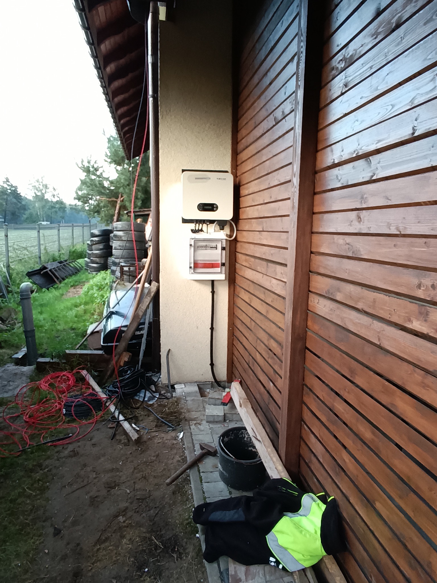 Biały falownik i licznik energii zamontowane na elewacji budynku, obok drewnianego ogrodzenia. Widoczne kable, narzędzia i odzież robocza na ziemi.