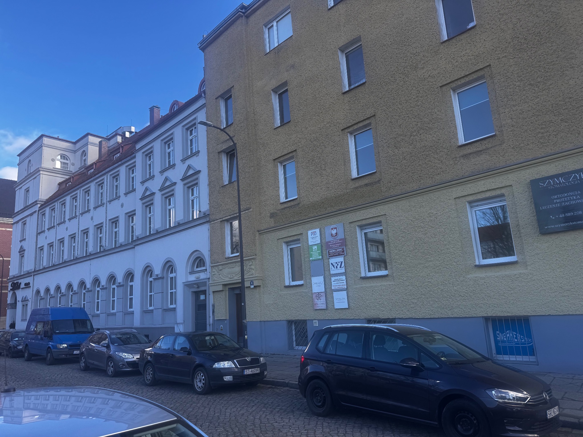 Ulica z zaparkowanymi samochodami przed budynkami, w tym jeden z szyldem stomatologicznym.