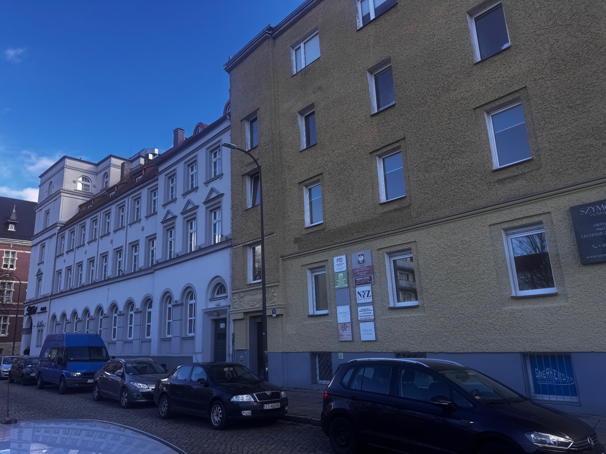 Ulica z zaparkowanymi samochodami przed budynkami, na jednym z nich szyldy kancelarii prawnych.