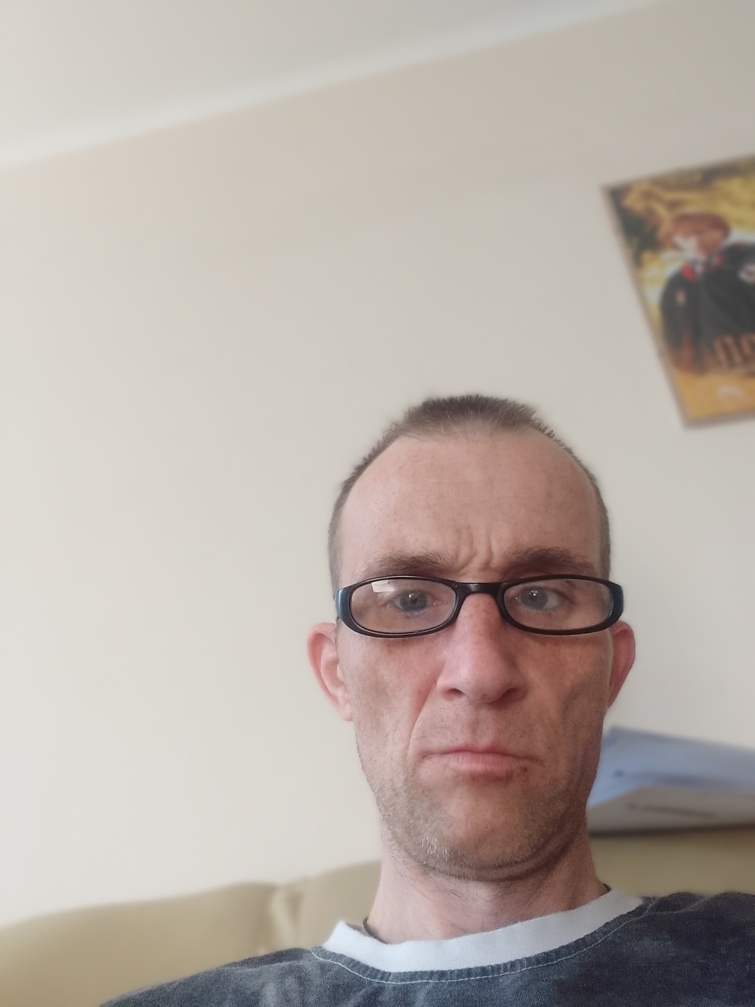 Portret mężczyzny w okularach z plakatem na ścianie w tle. Ujęcie z dołu, lekko zniekształcające perspektywę. Neutralny wyraz twarzy, codzienne ubranie.