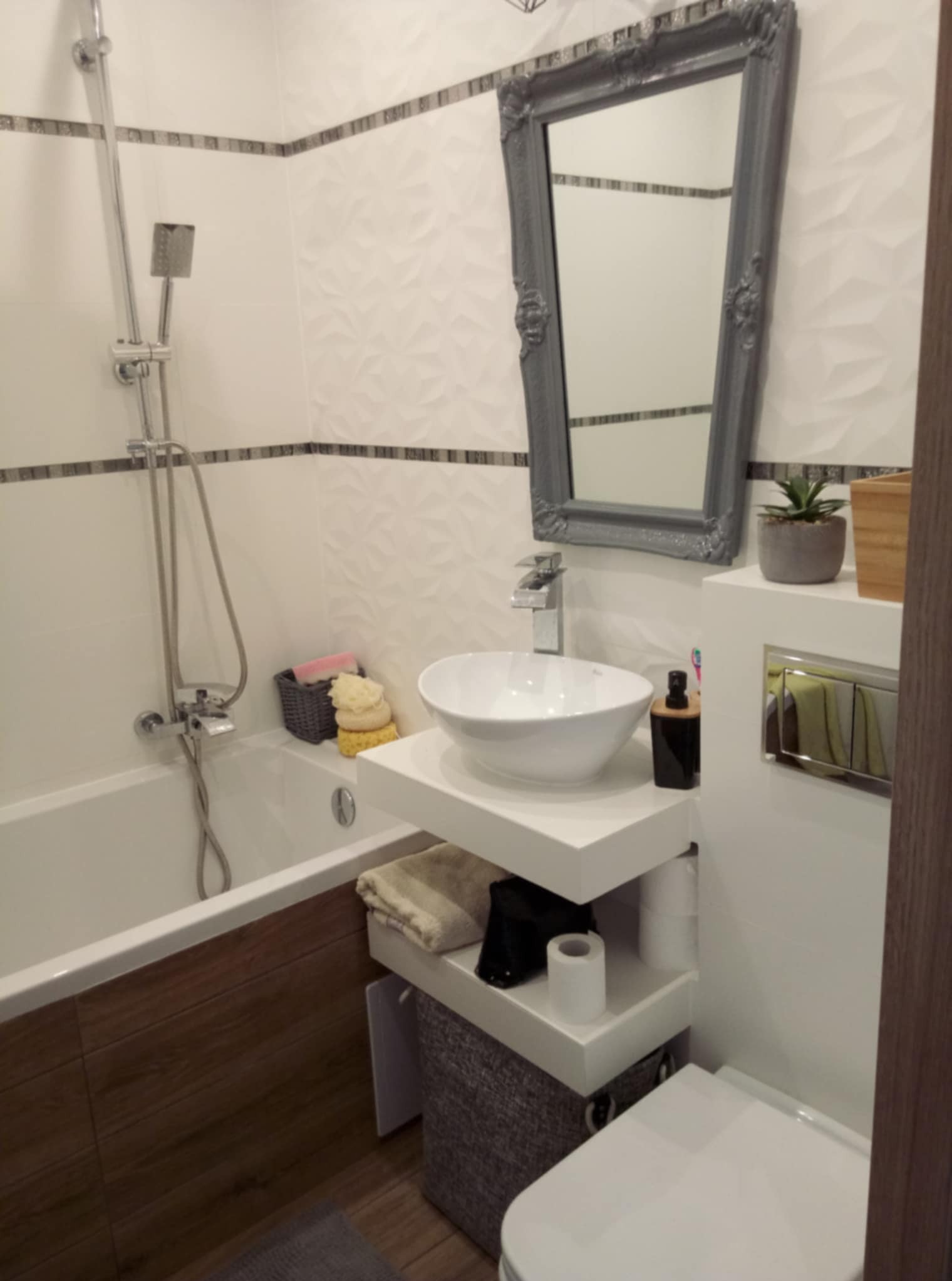 Nowoczesna łazienka z białą umywalką nablatową, szarą ramą lustra w stylu retro, mozaiką na ścianach i drewnopodobną obudową wanny.