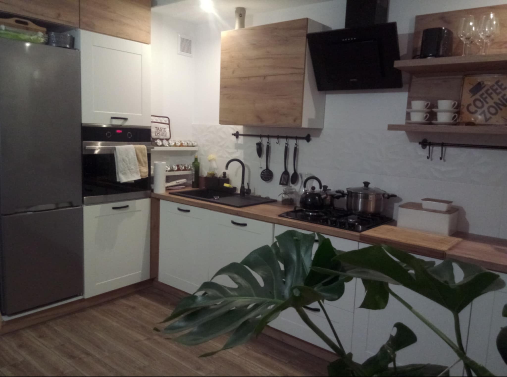 Nowoczesna kuchnia z białymi szafkami, drewnianymi akcentami i czarnym okapem, uzupełniona o roślinę Monstera w pierwszym planie.