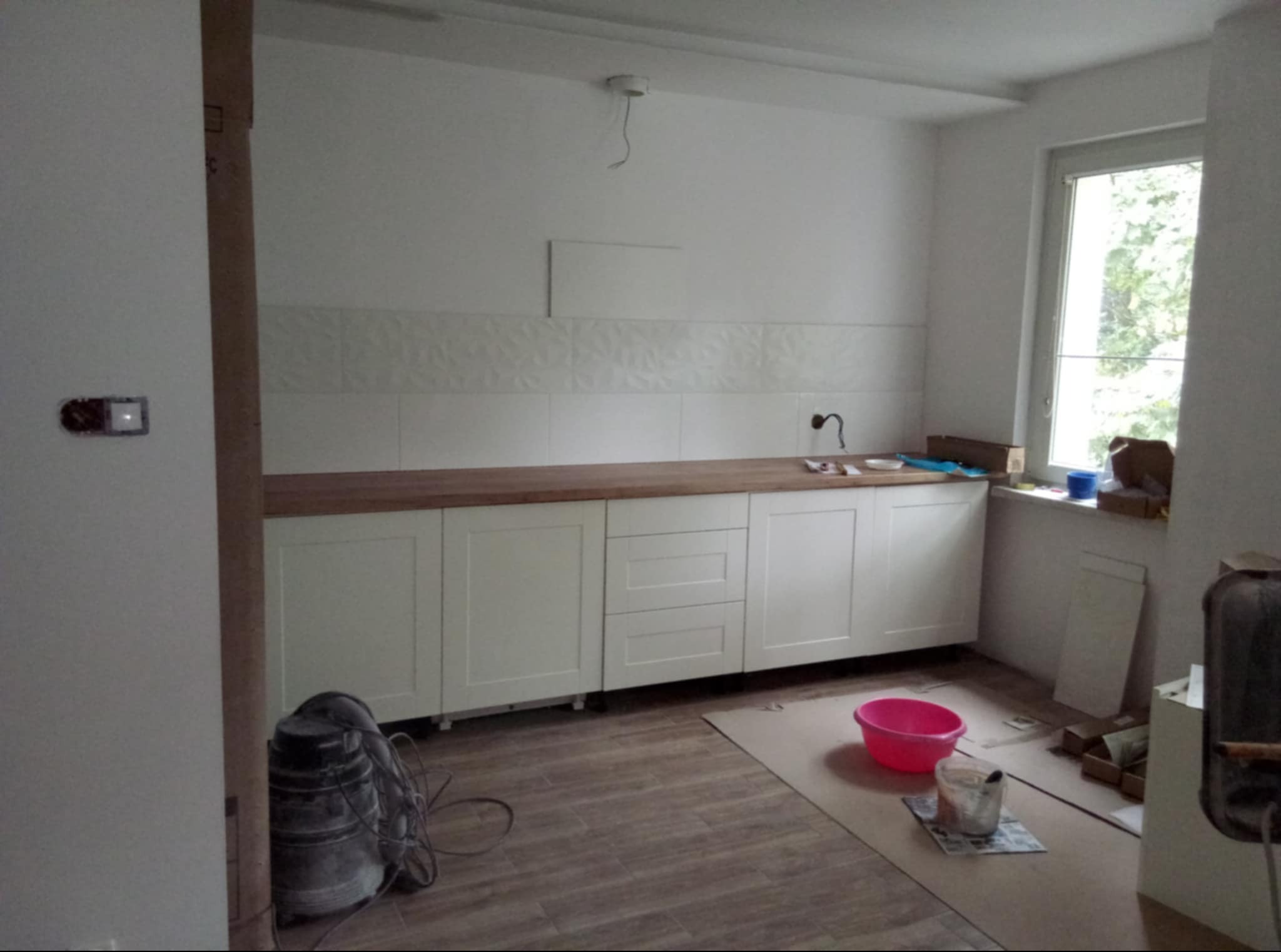 Wnętrze kuchni w trakcie prac wykończeniowych: białe szafki z drewnianym blatem, ułożone płytki dekoracyjne na ścianie, widoczne narzędzia i akcesoria malarskie, podłoga wyłożona panelami...