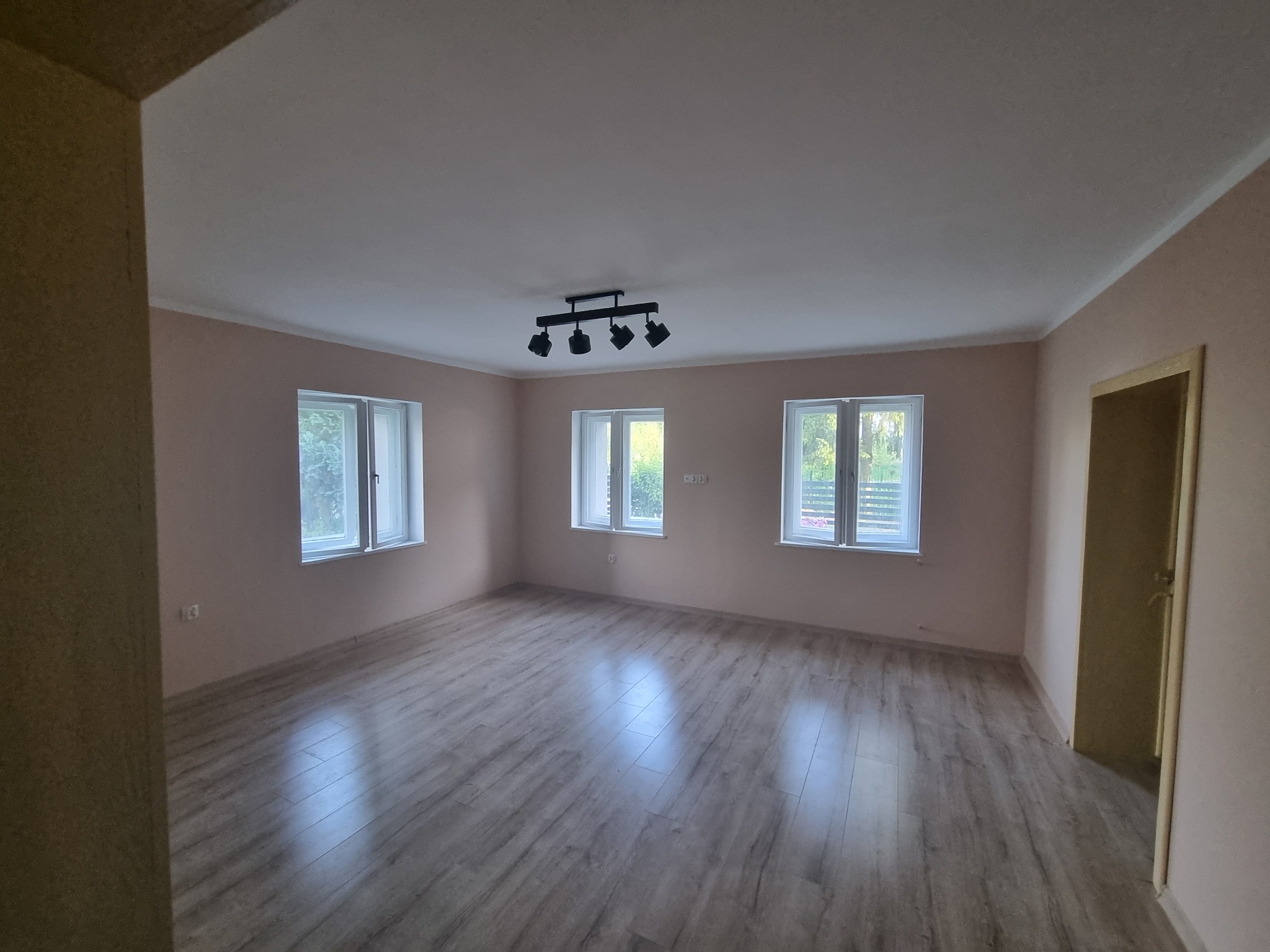 Pusty pokój po remoncie z panelami podłogowymi, trzema oknami i lampą sufitową z czterema reflektorami.