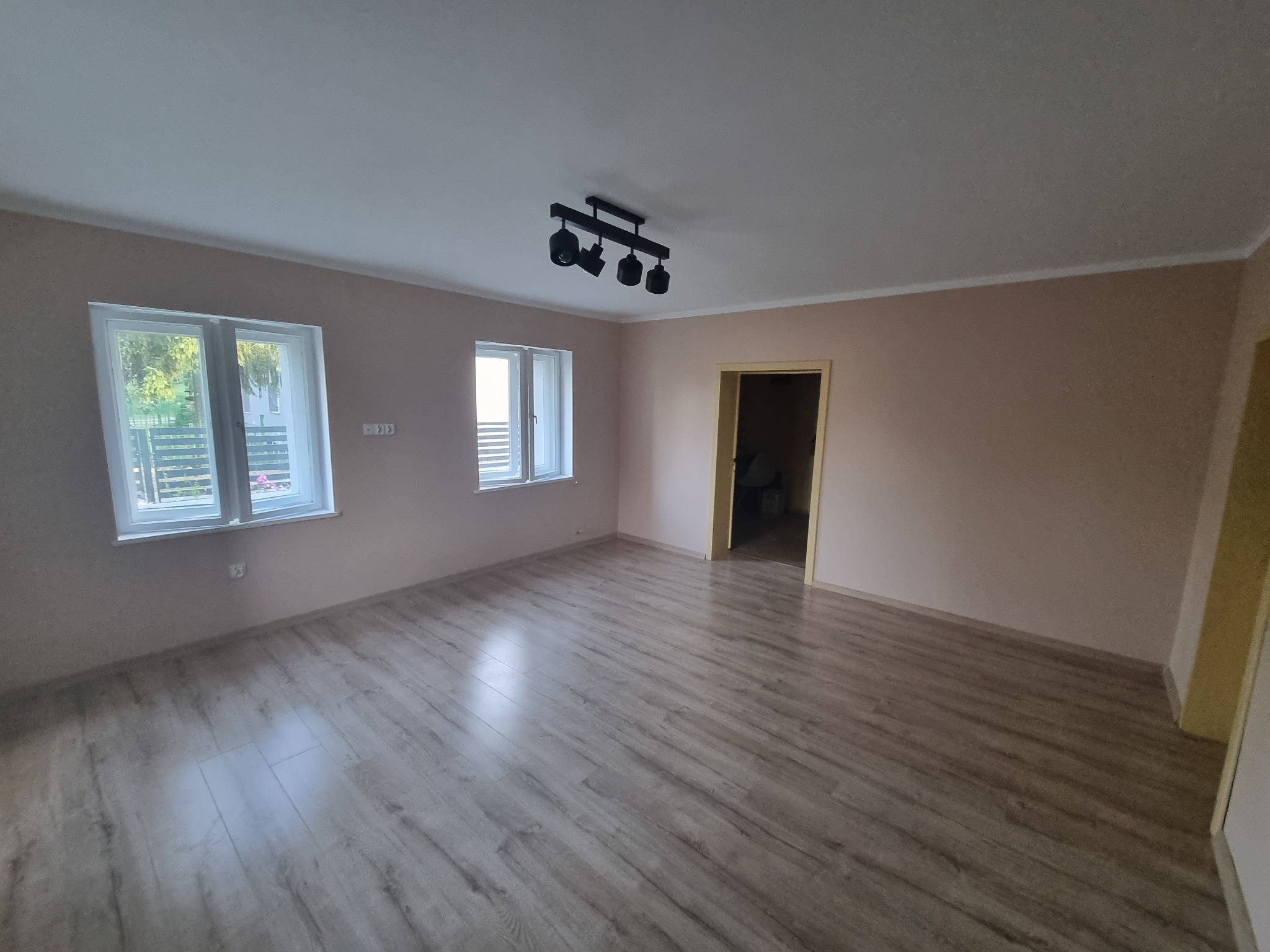 Pusty pokój po odświeżeniu, z panelami podłogowymi w odcieniach szarości, ścianami w kolorze beżowym, dwoma oknami z białymi ramami i czarną lampą sufitową z czterema reflektorami.