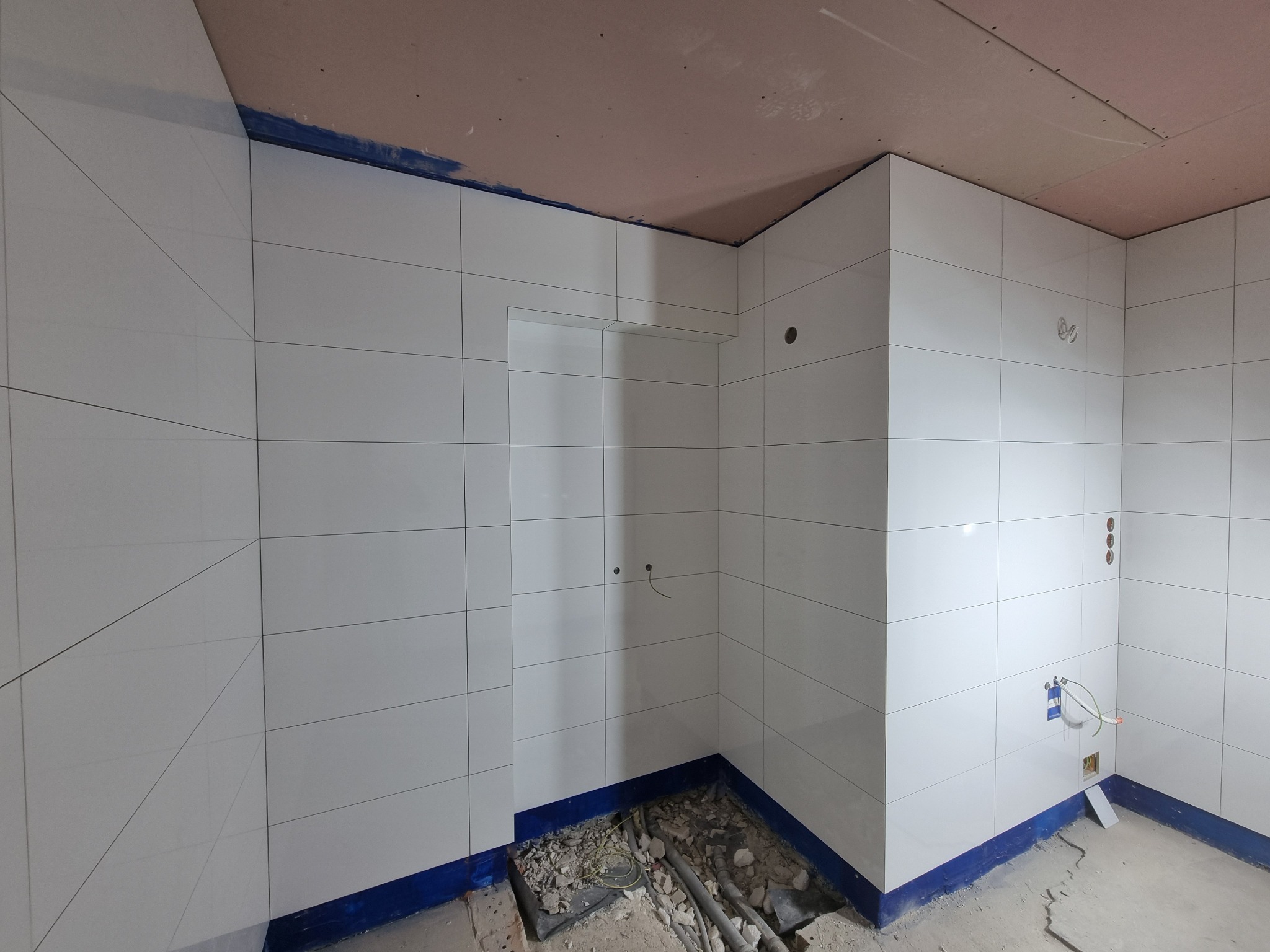 Białe płytki na ścianach łazienki w trakcie montażu, widoczne otwory na instalacje, niezagruntowany sufit z płyt kartonowo-gipsowych i nieukończona podłoga z fragmentami gruzu i rur.