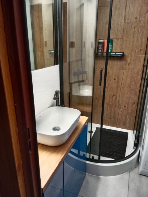 Nowoczesna łazienka z kabiną prysznicową z czarnymi ramami, umywalką nablatową na drewnianym blacie i płytkami imitującymi drewno na ścianie.