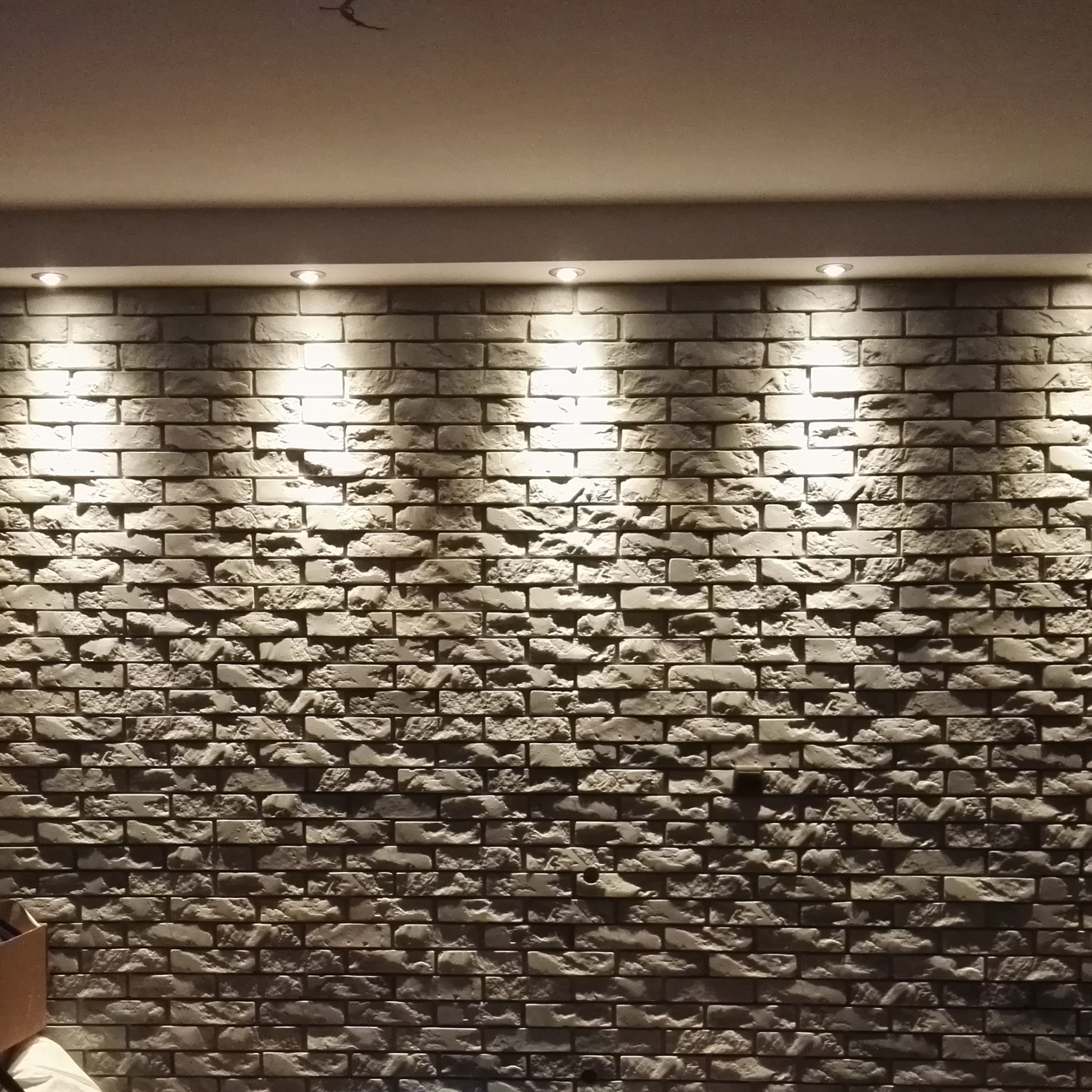 Ściana wyłożona dekoracyjną, szarą cegłą z nierówną fakturą, oświetlona trzema punktowymi lampami sufitowymi, tworzącymi grę światła i cienia.