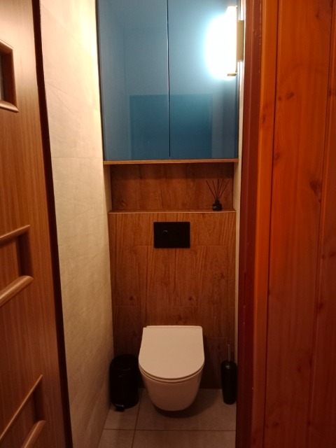 Nowoczesna toaleta wisząca w małej łazience z drewnianymi panelami ściennymi, niebieskimi szafkami wiszącymi i czarnymi akcesoriami, w tym szczotką do WC i koszem na śmieci.