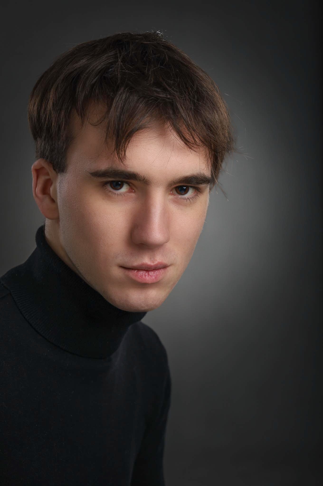 Portret młodego mężczyzny w czarnym golfie, oświetlonego studyjnie na szarym tle, z intensywnym spojrzeniem skierowanym wprost.