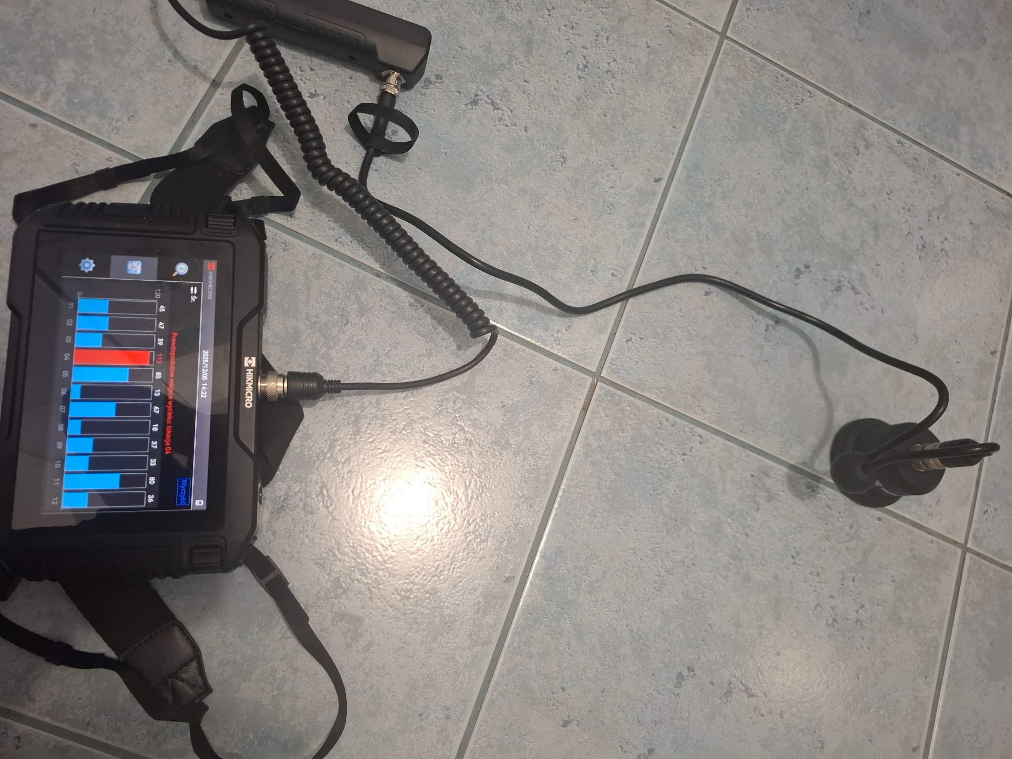 Urządzenie diagnostyczne HIKMICRO z ekranem prezentującym wykresy, prawdopodobnie używane do lokalizacji wycieków, leżące na płytkach podłogowych.