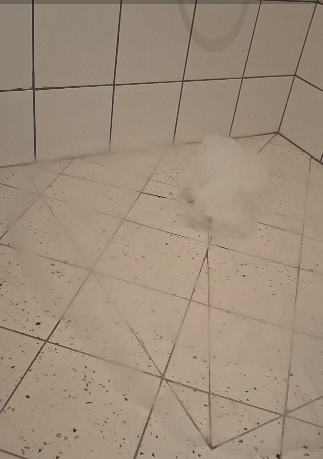 Białe płytki podłogowe i ścienne w łazience z widoczną parą/dymem, sugerujące test szczelności lub osuszanie po zalaniu. Ujęcie z niskiego kąta.