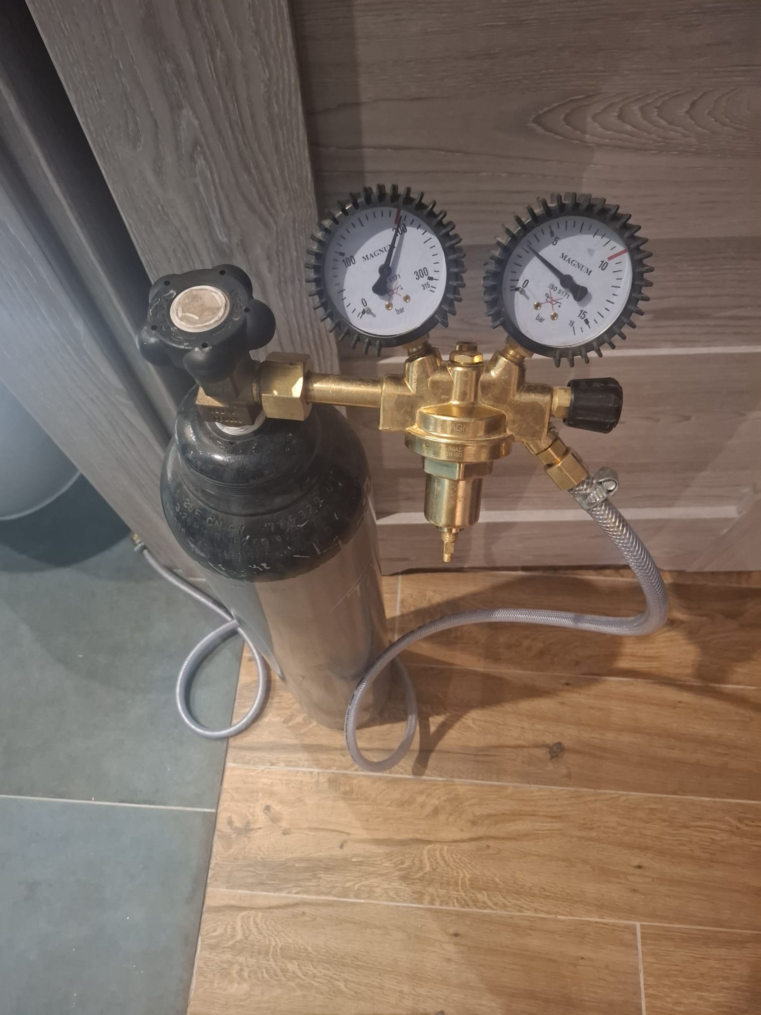 Butla z reduktorem ciśnienia i dwoma manometrami, ustawiona na podłodze z płytek imitujących drewno, na tle ściany z paneli. Widoczne detale złączek i węży.