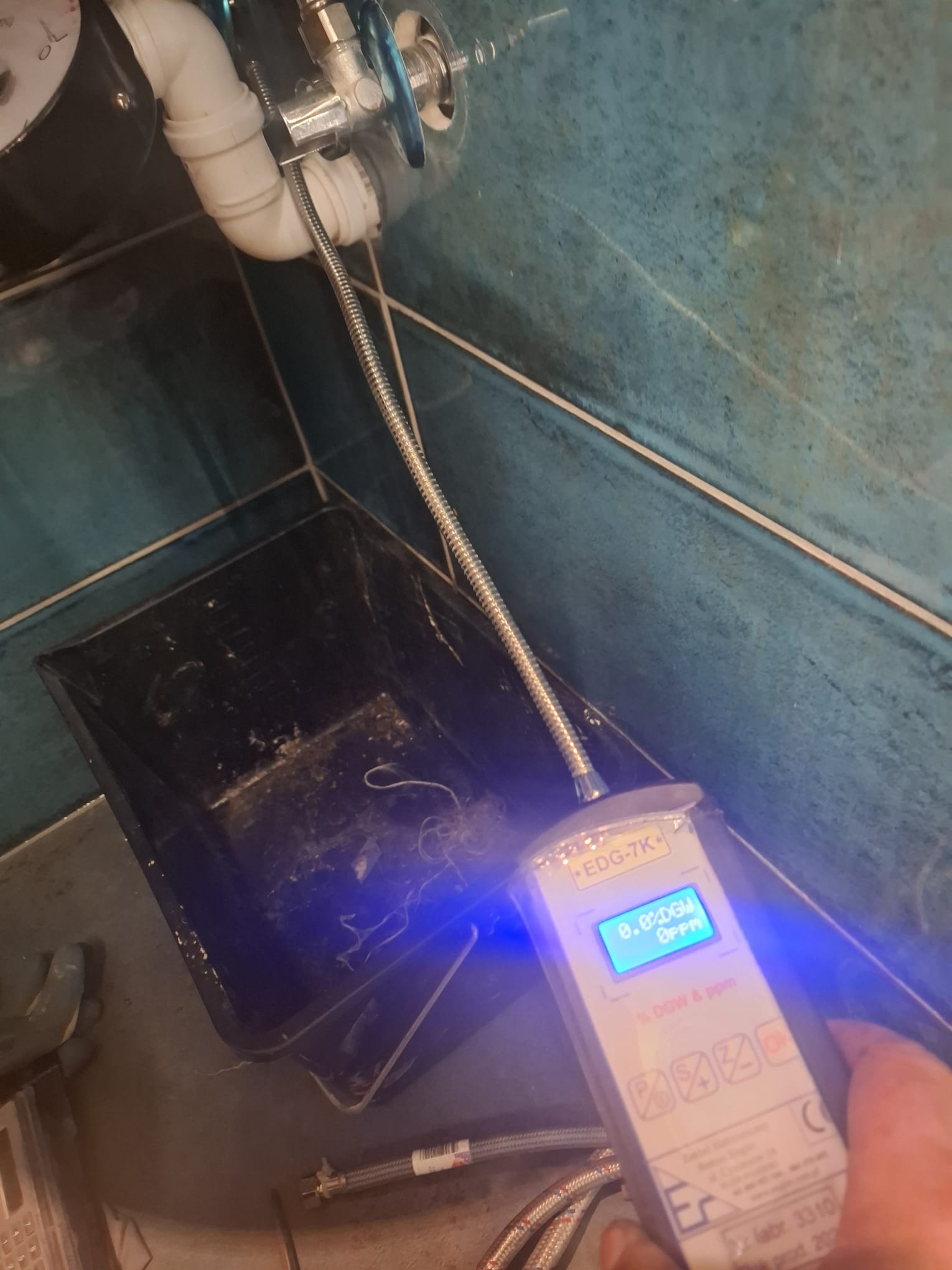 Pomiar wilgotności w odpływie umywalki za pomocą elektronicznego miernika EDG-7K. Widoczny syfon, rury i fragment ściany z niebieskich płytek.