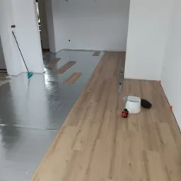 Układanie paneli podłogowych: widoczny podkład izolacyjny, częściowo ułożone panele w kolorze jasnego drewna, poziomica, laser krzyżowy, gumowy młotek i akcesoria.