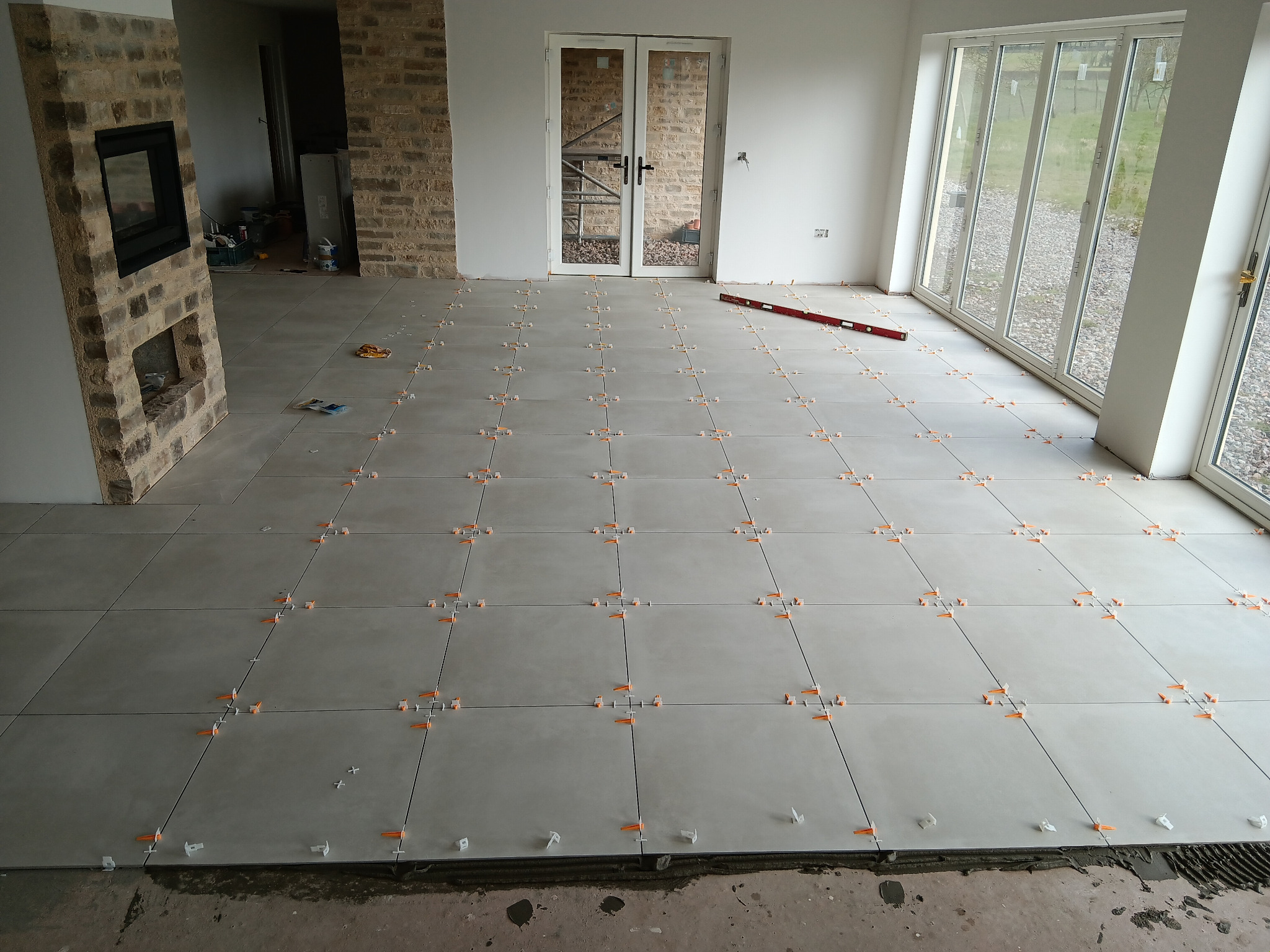 Układanie szarych płytek podłogowych z pomarańczowymi krzyżykami dystansowymi w przestronnym salonie z kominkiem z cegły i dużymi oknami tarasowymi.