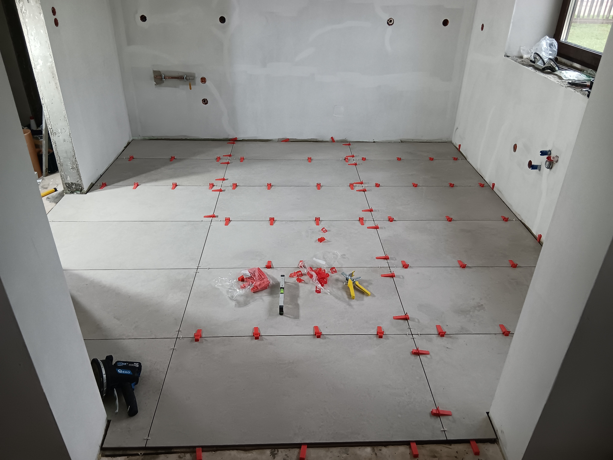 Szare płytki podłogowe w trakcie układania, z czerwonymi klipsami poziomującymi, widoczne narzędzia glazurnicze i poziomica, surowe ściany przygotowane do dalszych prac wykończeniowych.