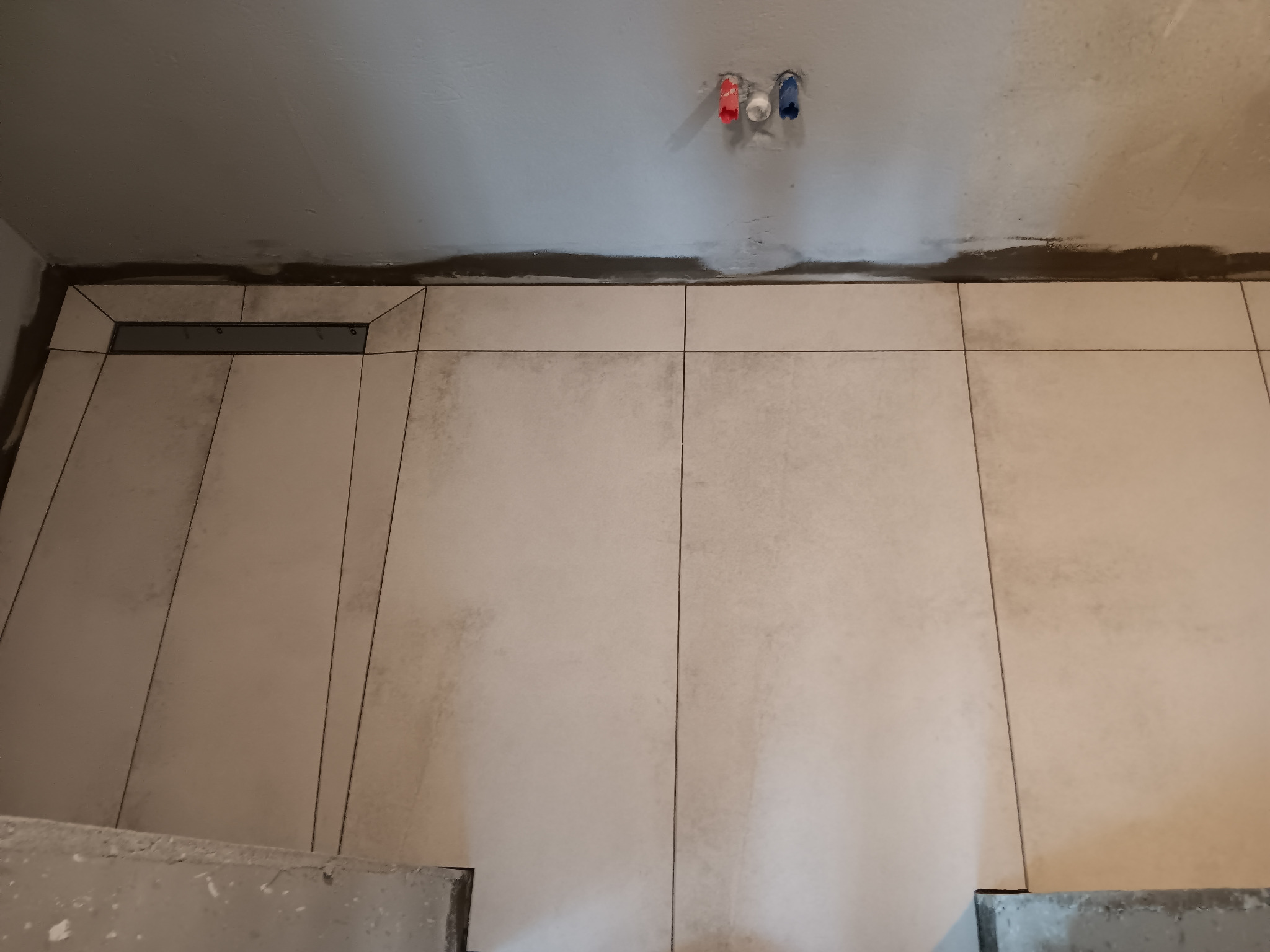 Ściana łazienki w trakcie wykańczania, widoczne jasne płytki ceramiczne, odpływ liniowy w podłodze, wystające rury instalacji wodnej ze ściany.