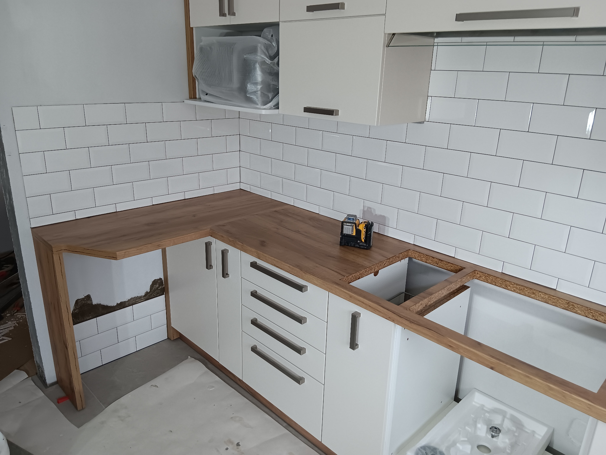 Montaż blatu drewnopodobnego w kuchni z białymi szafkami i białymi płytkami metra, widoczny laser krzyżowy i fragmenty niezakończonego układania płytek.