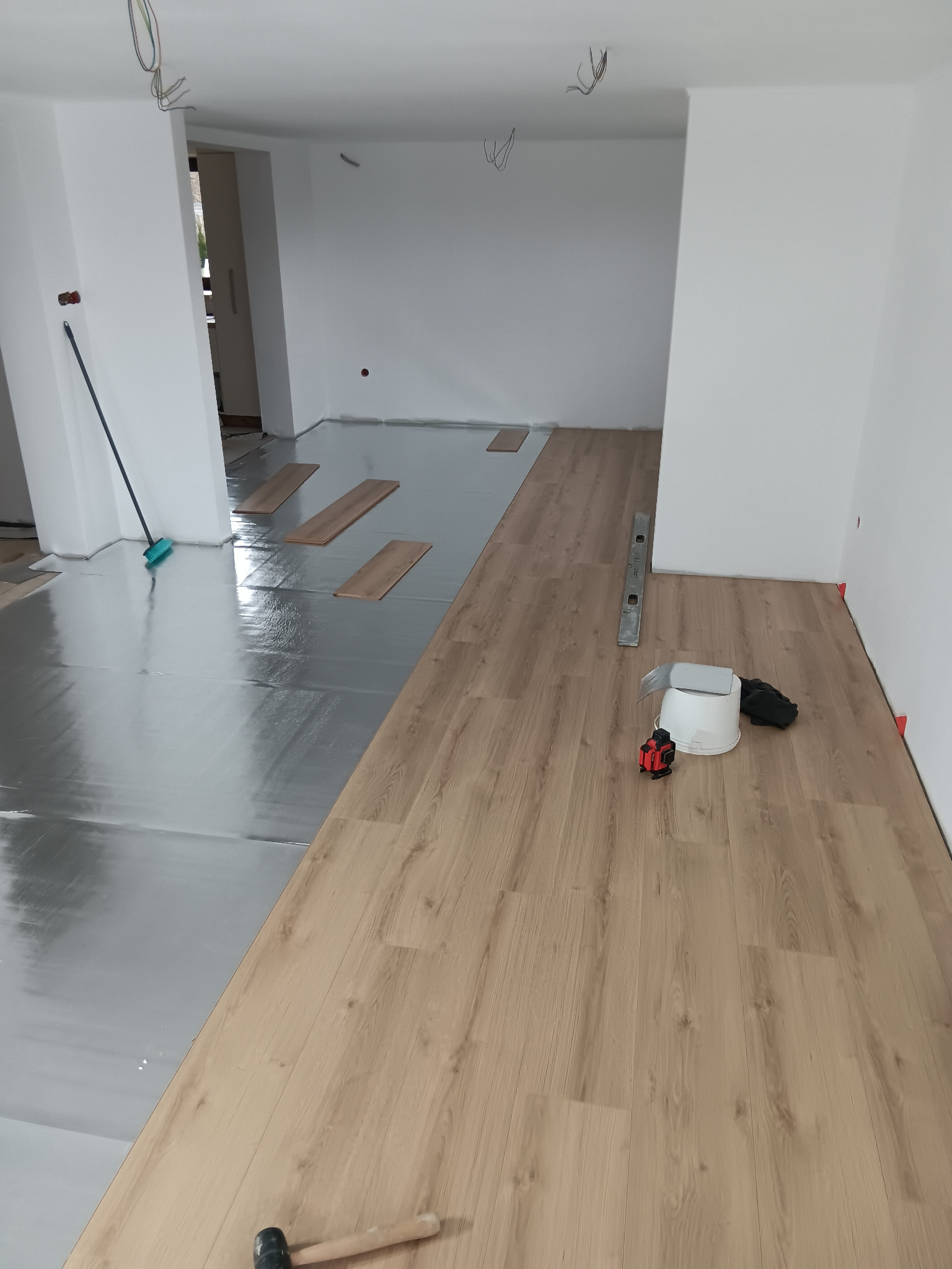 Układanie paneli podłogowych: widoczny podkład izolacyjny, częściowo ułożone panele w kolorze jasnego drewna, poziomica, laser krzyżowy, gumowy młotek i akcesoria.