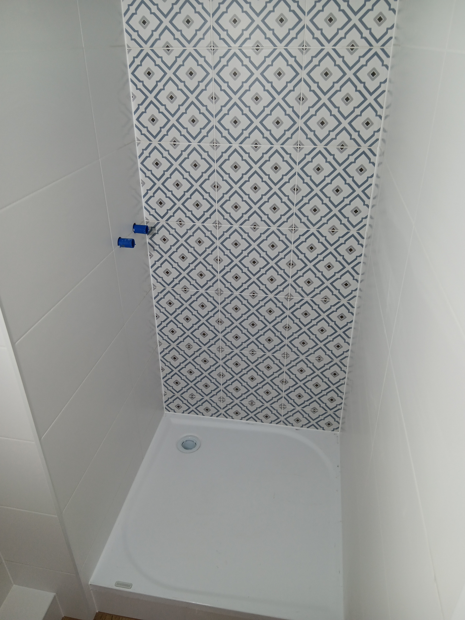 Biały brodzik z odpływem i fragment ściany wyłożonej dekoracyjnymi, niebiesko-białymi płytkami w geometryczne wzory. Widoczne niebieskie elementy instalacji.