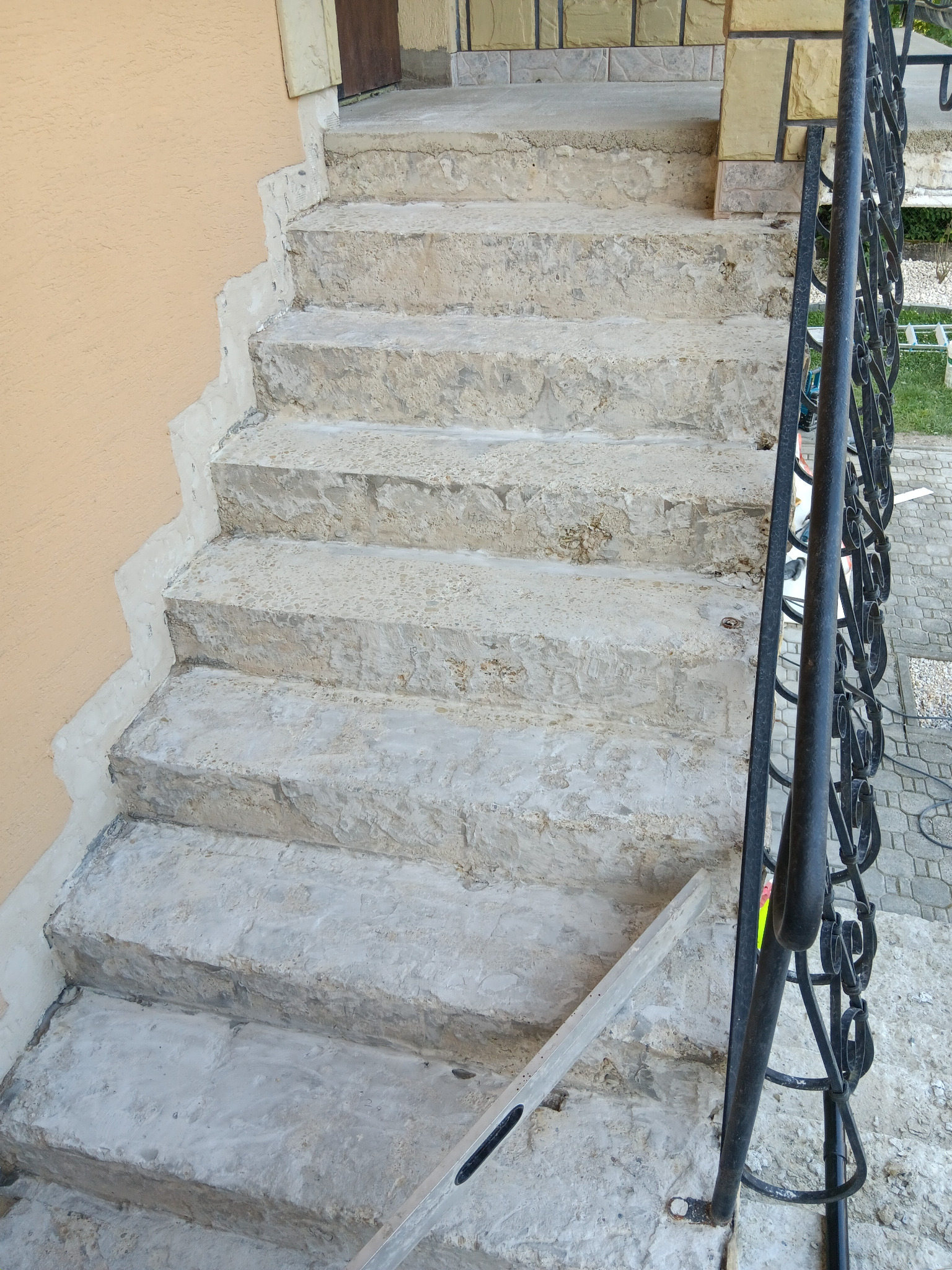 Schody zewnętrzne z betonu w trakcie naprawy, widoczne ślady zaprawy i poziomica, balustrada z kutego żelaza. Prace remontowe na zewnątrz budynku.