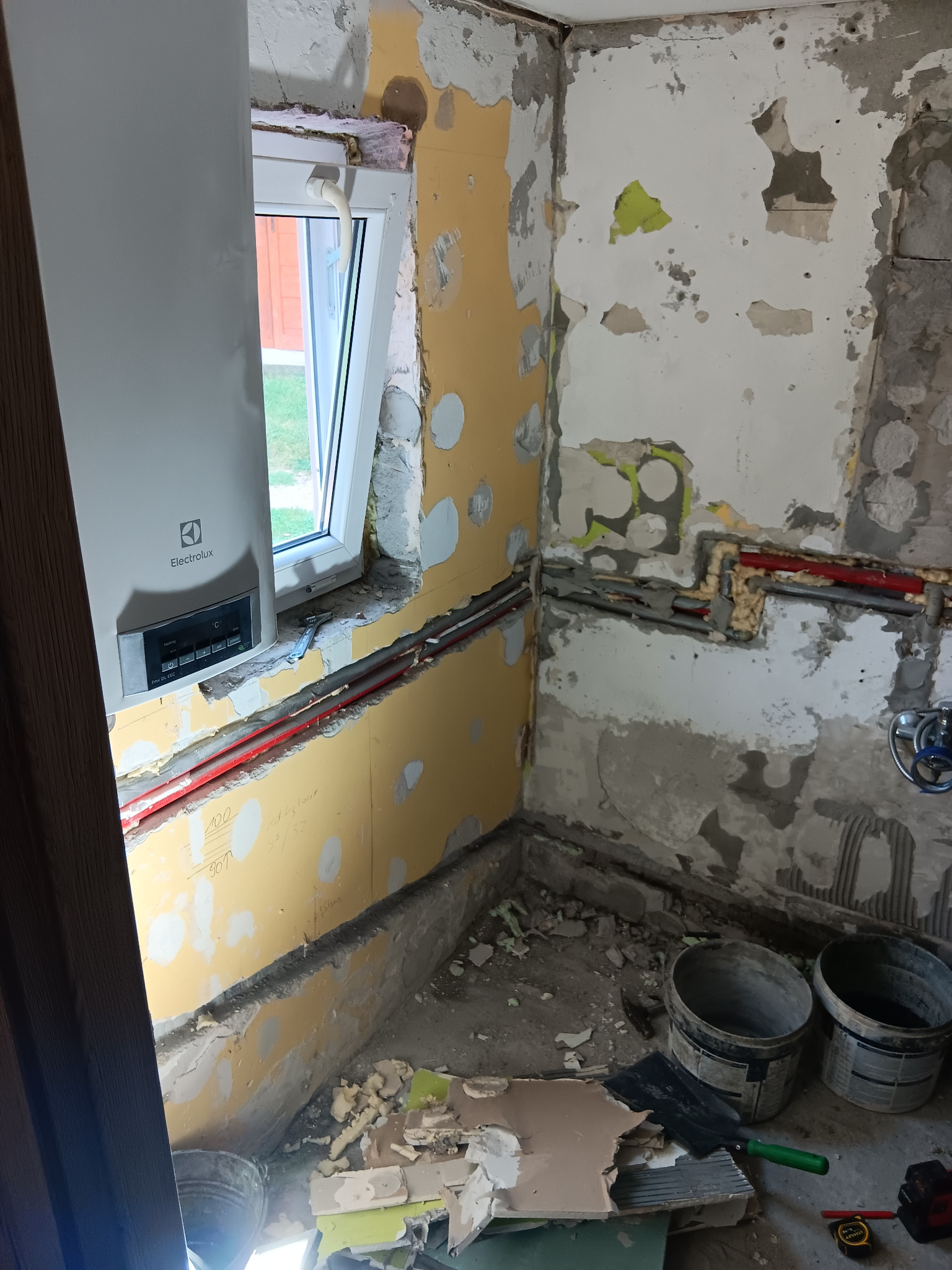 Stan surowy łazienki w trakcie prac: odsłonięte rury, przygotowane ściany pod płytki, nowy bojler elektryczny Electrolux przy oknie.