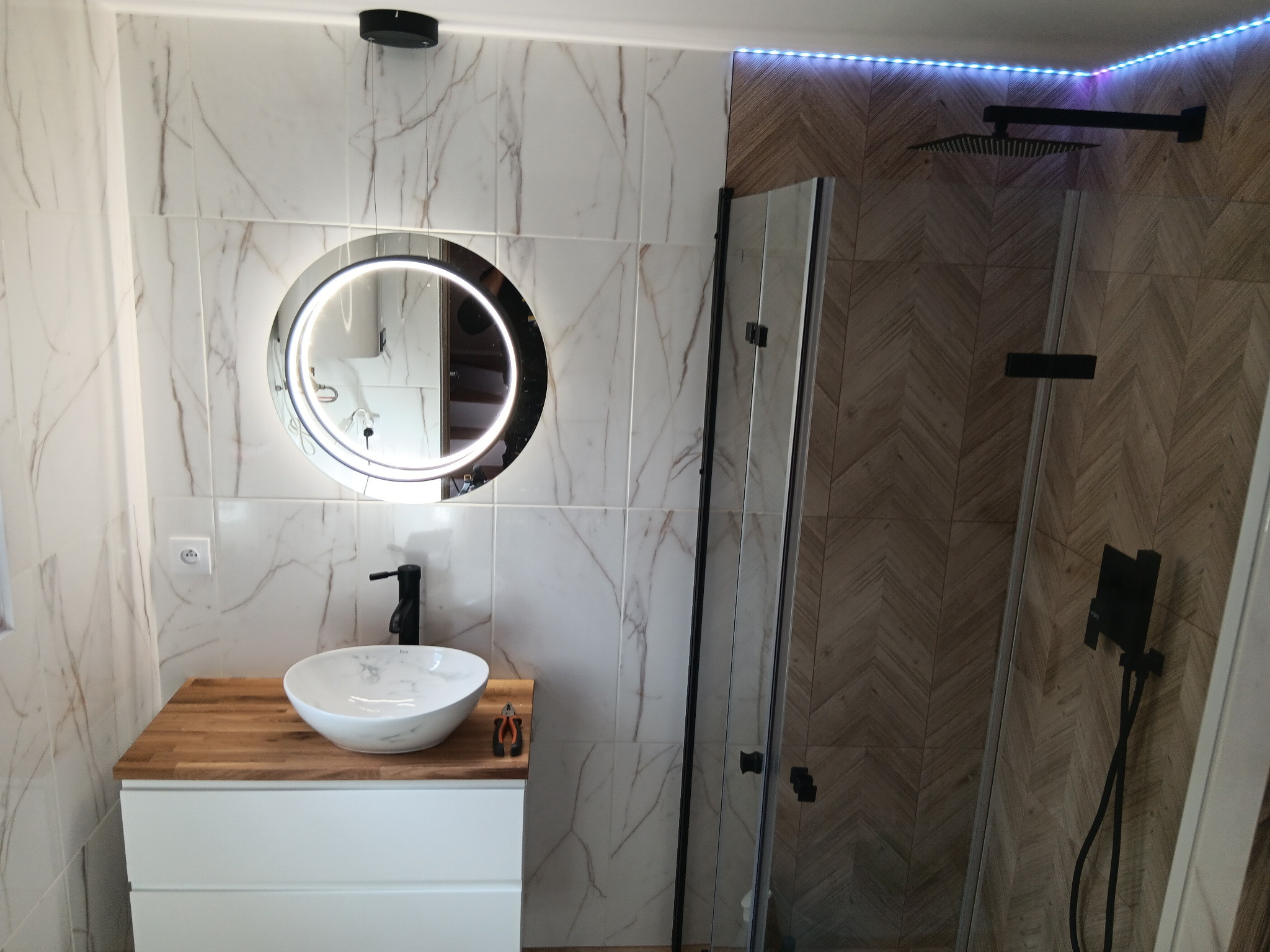 Wykończona łazienka z okrągłym lustrem LED, białą umywalką na drewnianym blacie i kabiną prysznicową z czarną armaturą. Na blacie leżą kombinerki.
