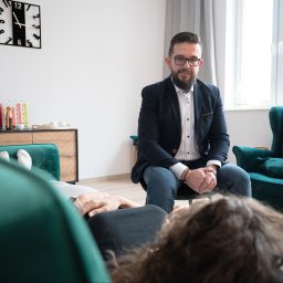 Zrozumieć Siebie - Centrum Hipnoterapii i Uwalniania Emocji - Mężczyzna w okularach i marynarce siedzi naprzeciwko kobiety leżącej na zielonej sofie w jasnym gabinecie z zegarem na ścianie.