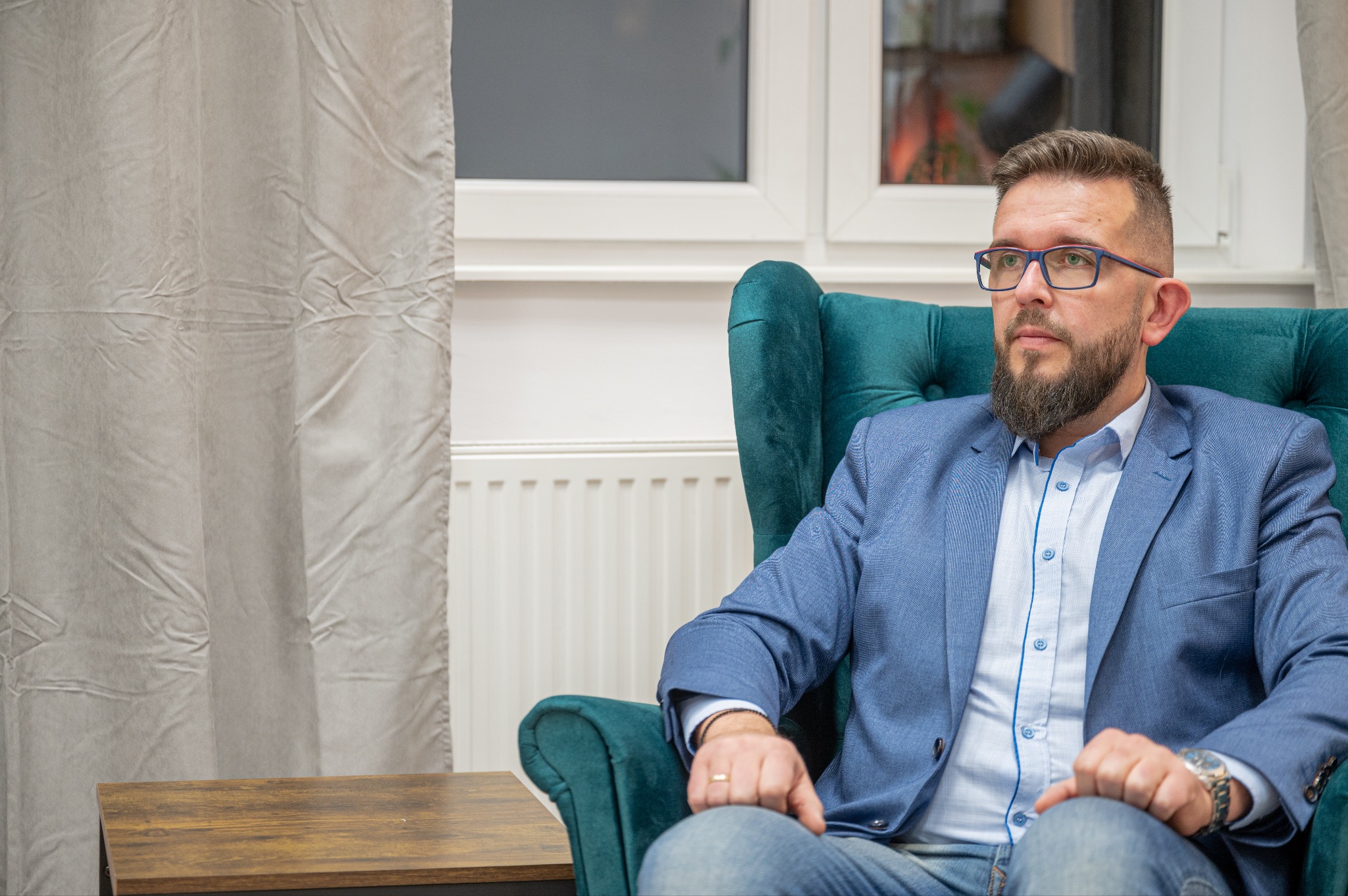 Mężczyzna w okularach i niebieskiej marynarce siedzi w zielonym fotelu, spoglądając w dal, w tle zasłona i okno.