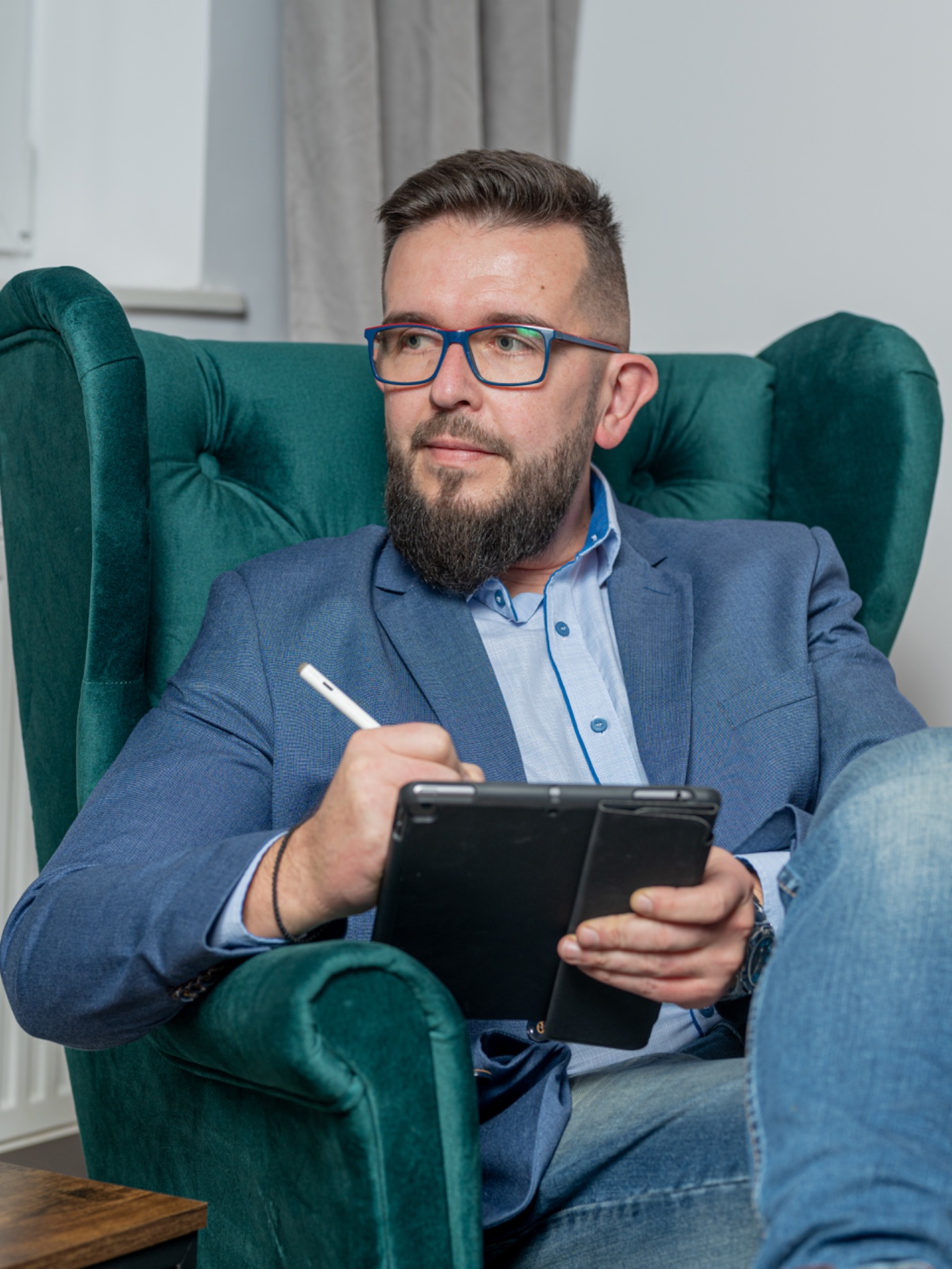 Mężczyzna z brodą w niebieskiej marynarce i okularach siedzi w zielonym fotelu, trzyma tablet i rysik, spoglądając w bok.