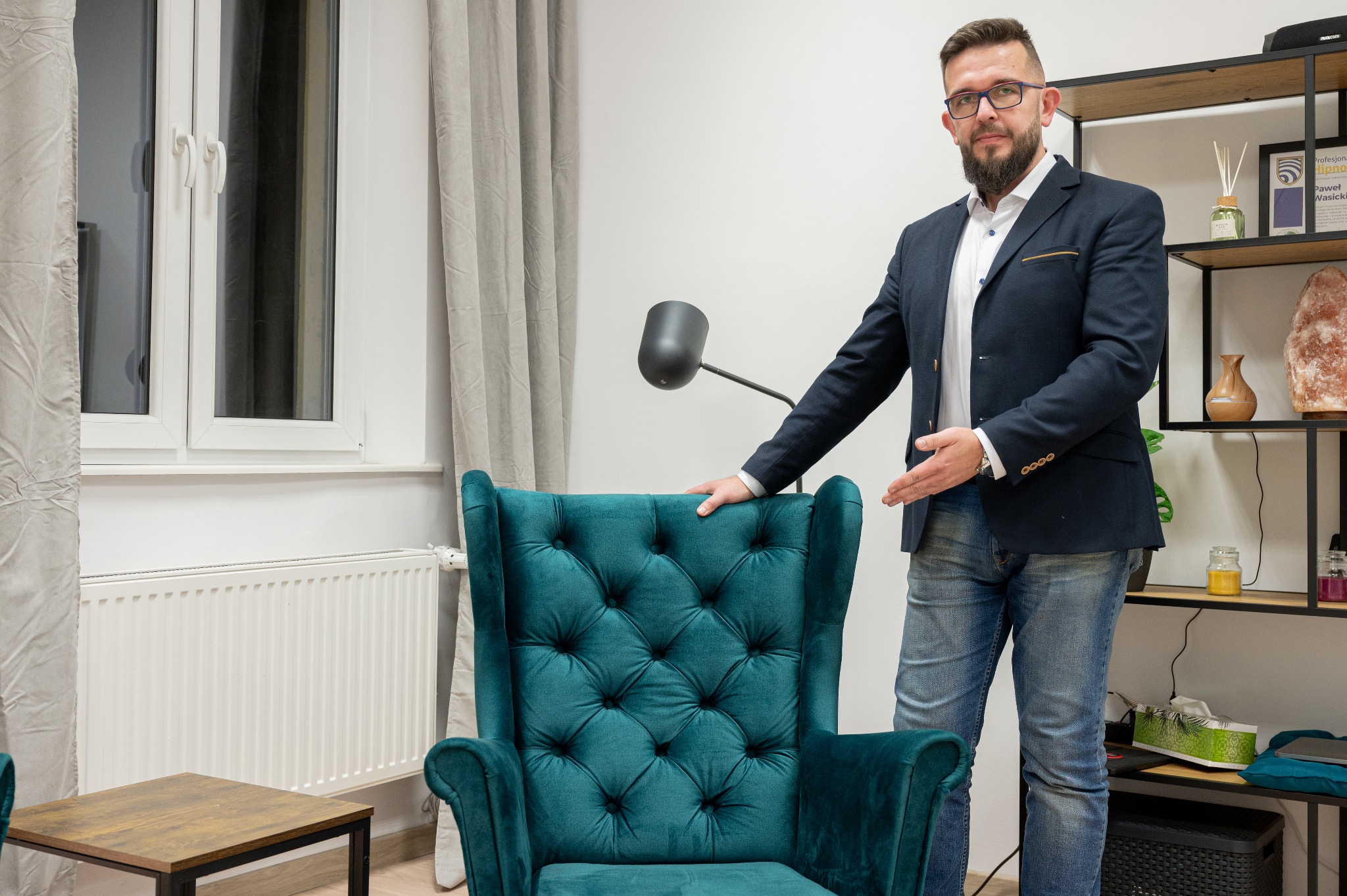 Mężczyzna w okularach i marynarce prezentuje turkusowy fotel typu uszak w gabinecie z półką, lampą i oknem.