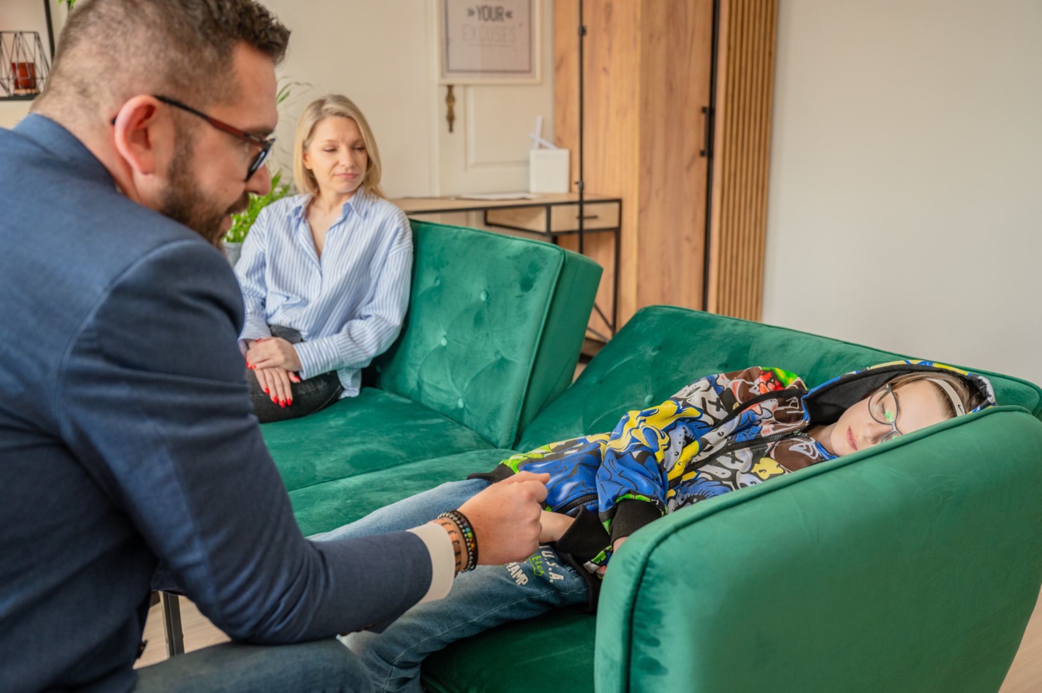 Sesja terapeutyczna: mężczyzna w garniturze siedzi obok leżącej na zielonej sofie osoby, w tle kobieta. Jasne wnętrze, naturalne światło.