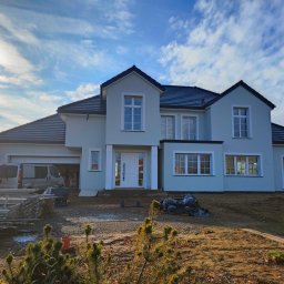 Elewacje-ciepły dom - Jasny, dwukondygnacyjny dom z ciemnym dachem po renowacji elewacji, z widocznym zaparkowanym vanem i elementami rusztowania. Błękitne niebo z chmurami w tle.