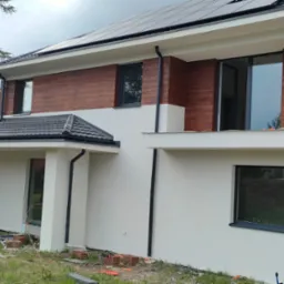 Nowoczesny dom z elewacją w kolorze bieli i drewna, okna w ciemnych ramach, panele słoneczne na dachu. Widoczne materiały budowlane.