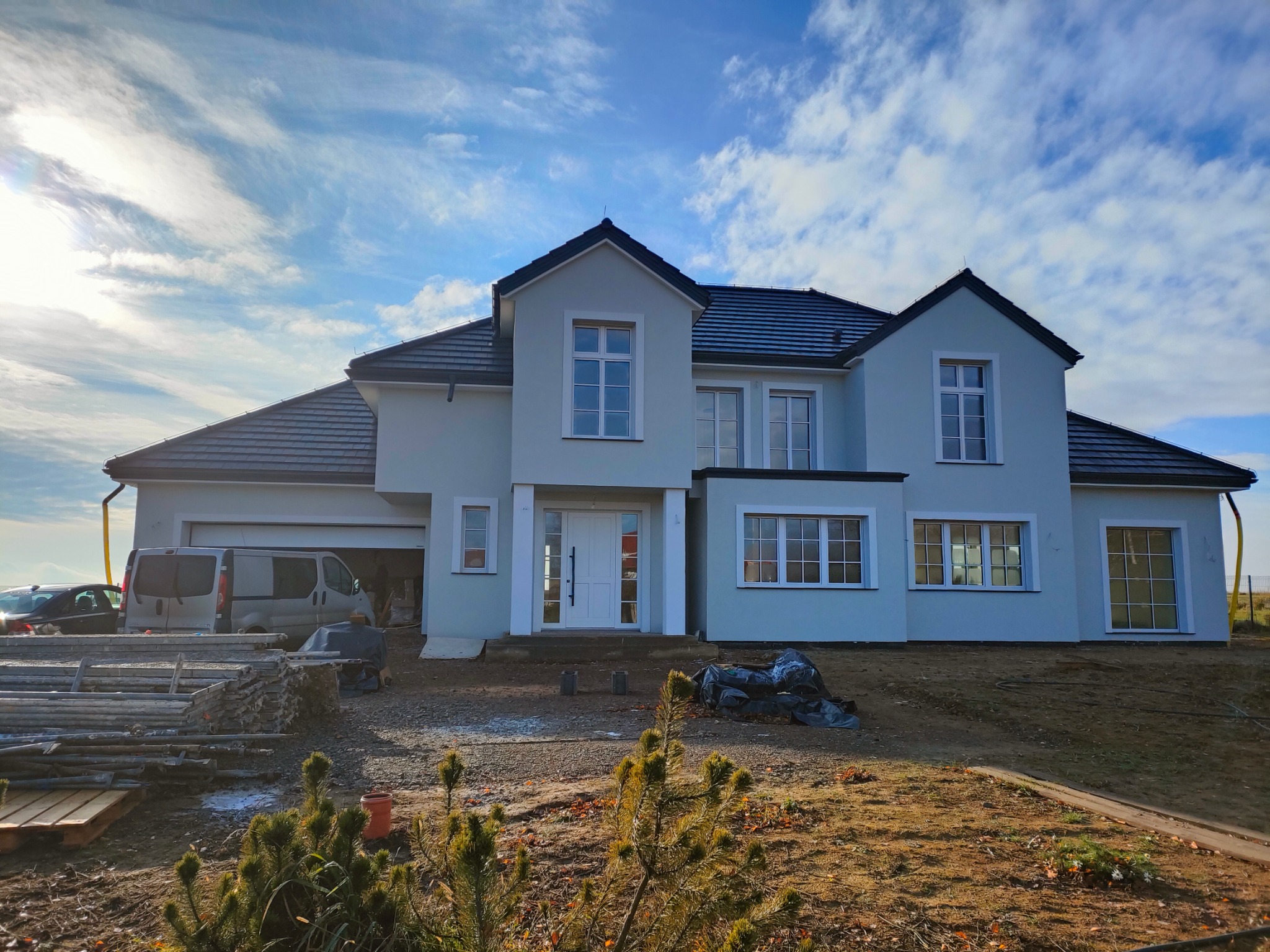 Jasny, dwukondygnacyjny dom z ciemnym dachem po renowacji elewacji, z widocznym zaparkowanym vanem i elementami rusztowania. Błękitne niebo z chmurami w tle.