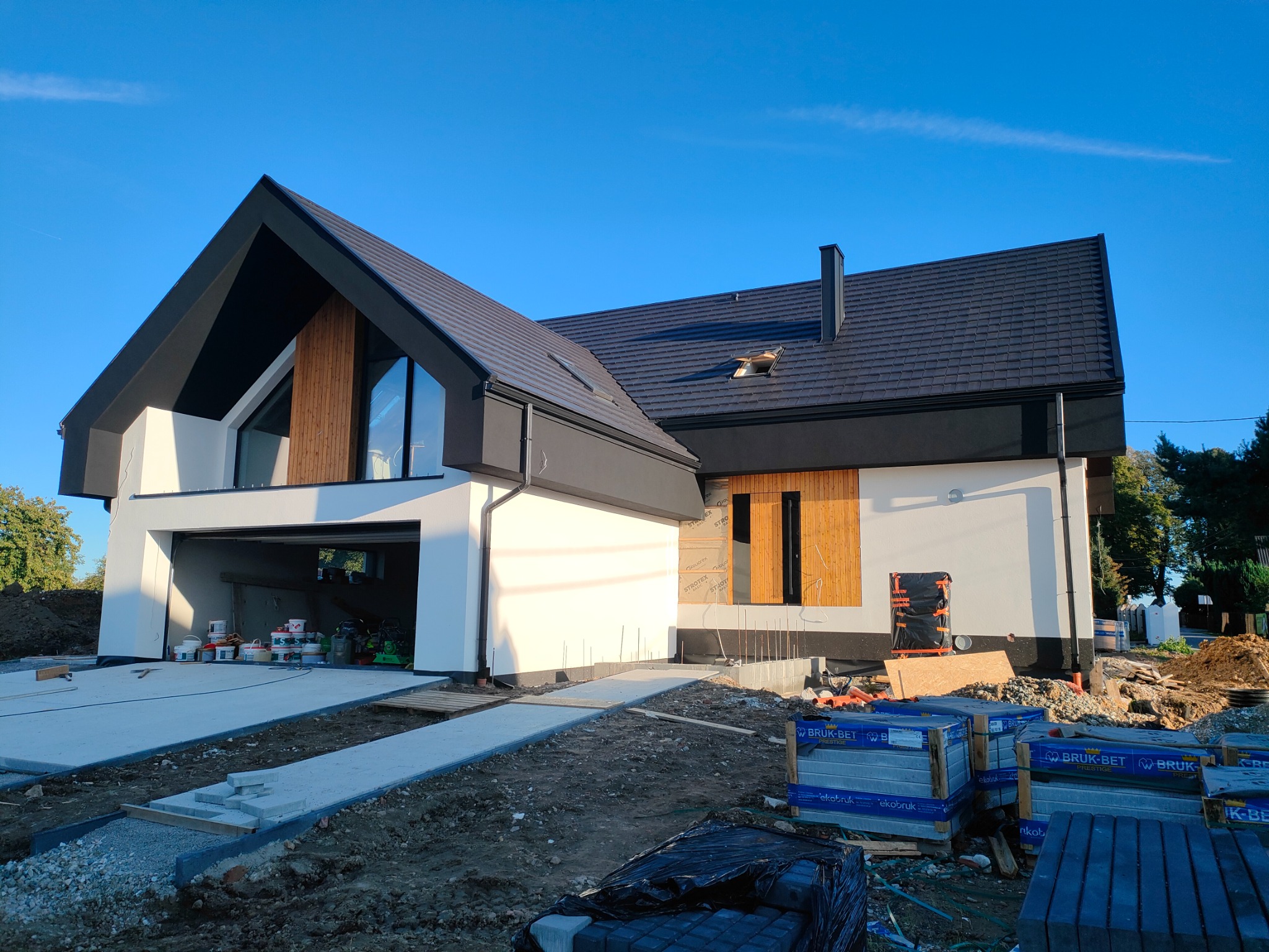 Nowoczesny dom z białą elewacją i czarnym dachem, widoczny w trakcie prac wykończeniowych. Na pierwszym planie kostka brukowa w opakowaniach. Błękitne niebo.