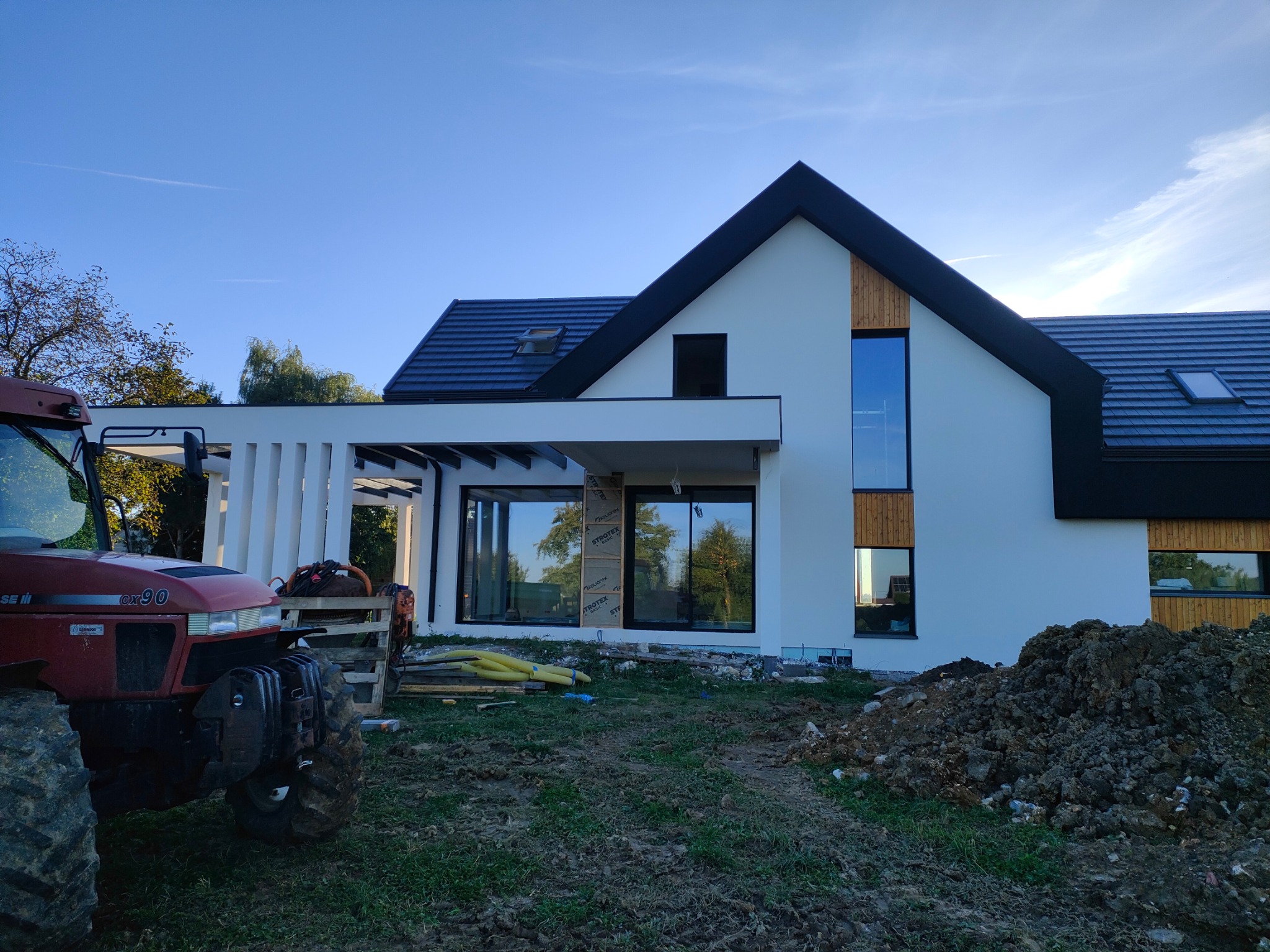Nowoczesny, biały dom z czarnym dachem w trakcie ocieplania. Widoczne płyty izolacyjne Strotex i traktor Case CX90. Świeże nasadzenia i ziemia.