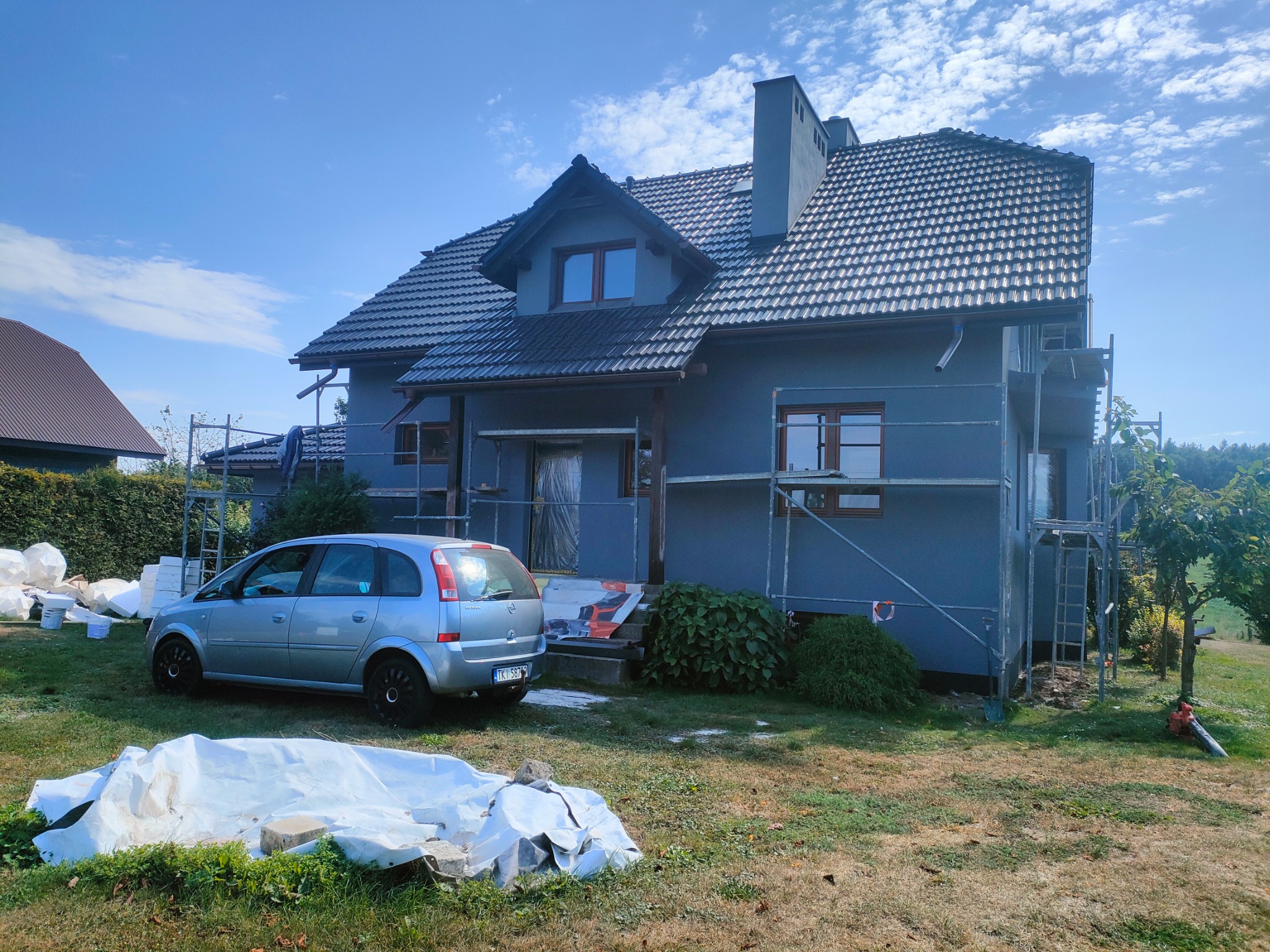 Dom jednorodzinny w trakcie prac elewacyjnych, widoczne rusztowania, materiały budowlane i samochód osobowy na posesji w słoneczny dzień.