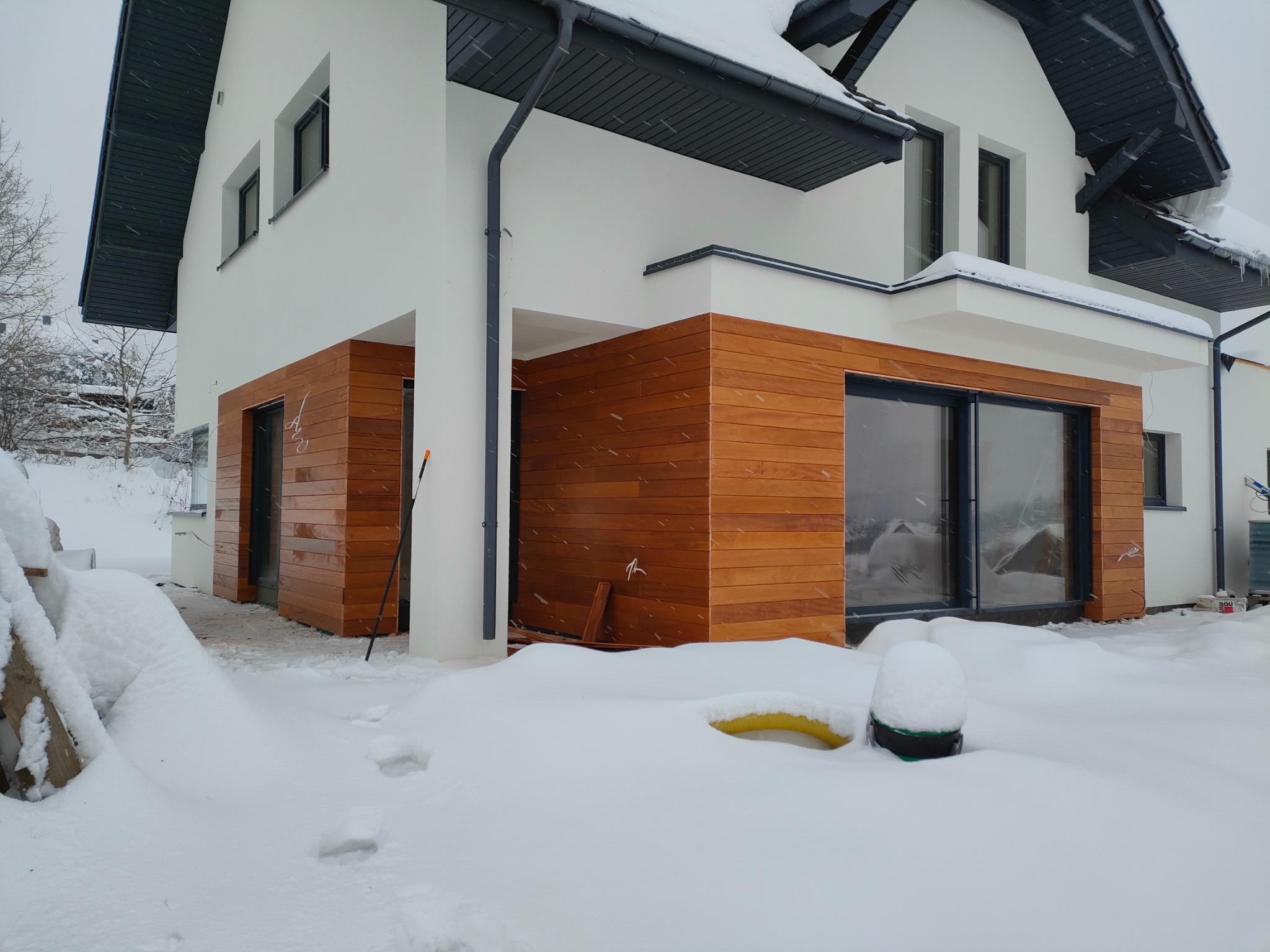 Nowoczesny dom z elewacją z jasnego drewna i białymi ścianami w zimowej scenerii. Śnieg pokrywa teren wokół budynku, a na dachu widać nawisy śnieżne.
