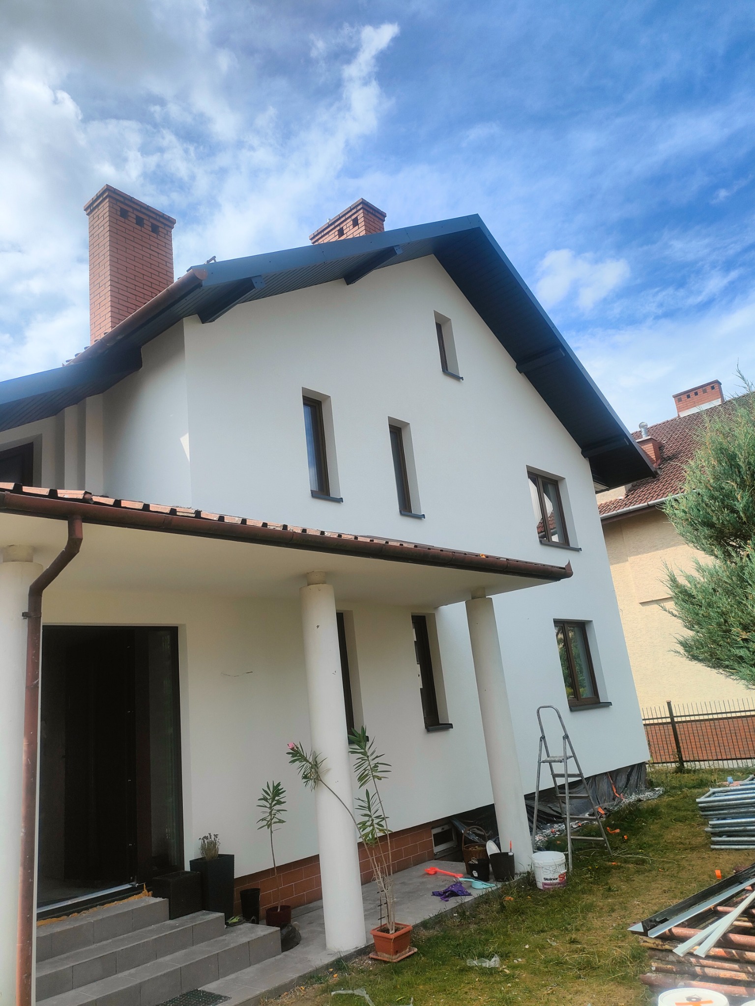 Dom jednorodzinny po renowacji elewacji. Biała fasada, ciemna stolarka okienna, ceglany komin. Widoczne rusztowania i drabina, co sugeruje niedawne prace.